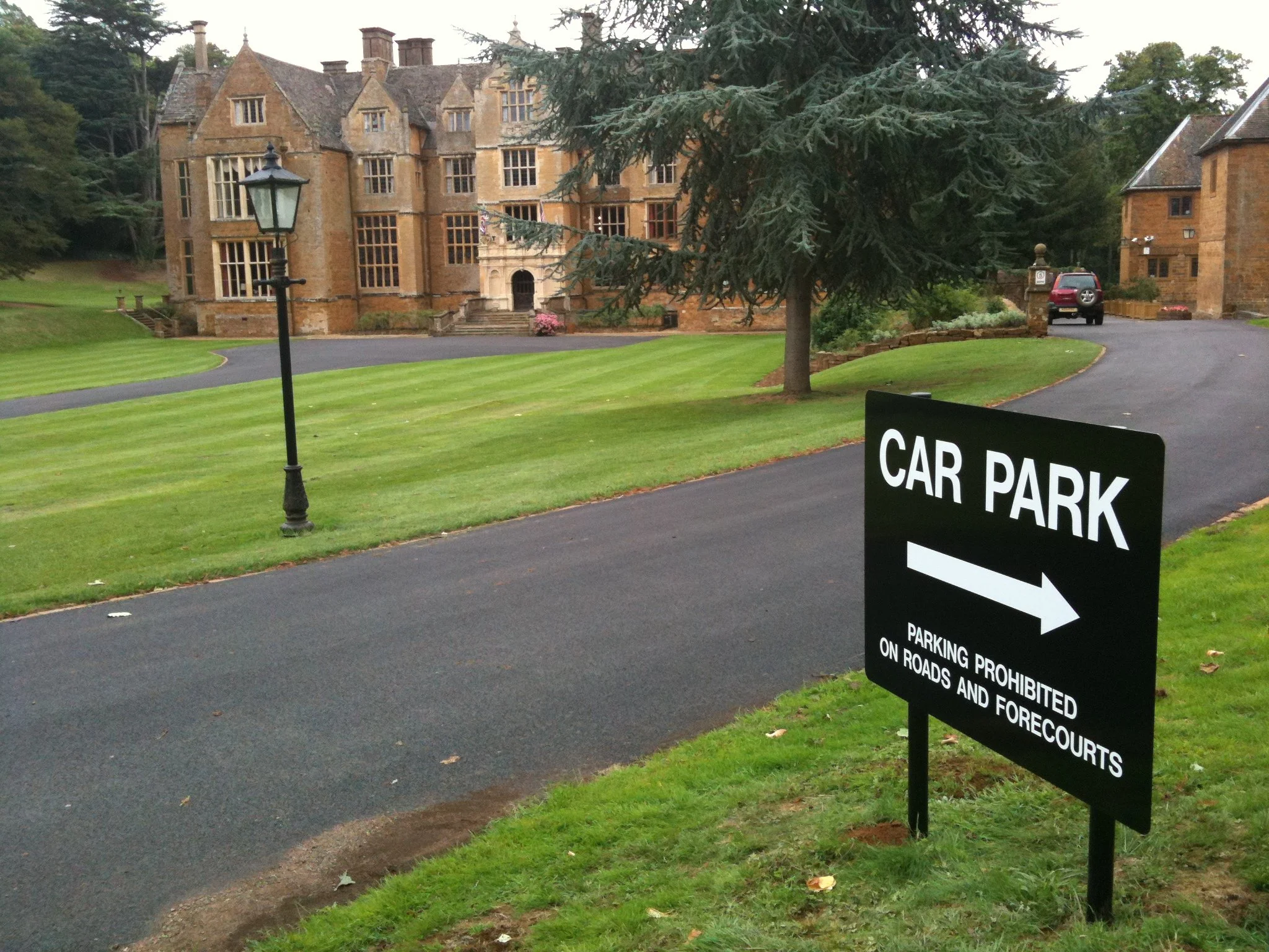 A gravel driveway leading to a large, historic stone mansion with multiple gables and large windows, surrounded by a well-maintained lawn with trees. A black streetlamp stands on the lawn. A black sign with white text and arrow indicates the direction to the car park, with a note that parking is prohibited on roads and forecourts. A car is parked on a curved driveway to the right.