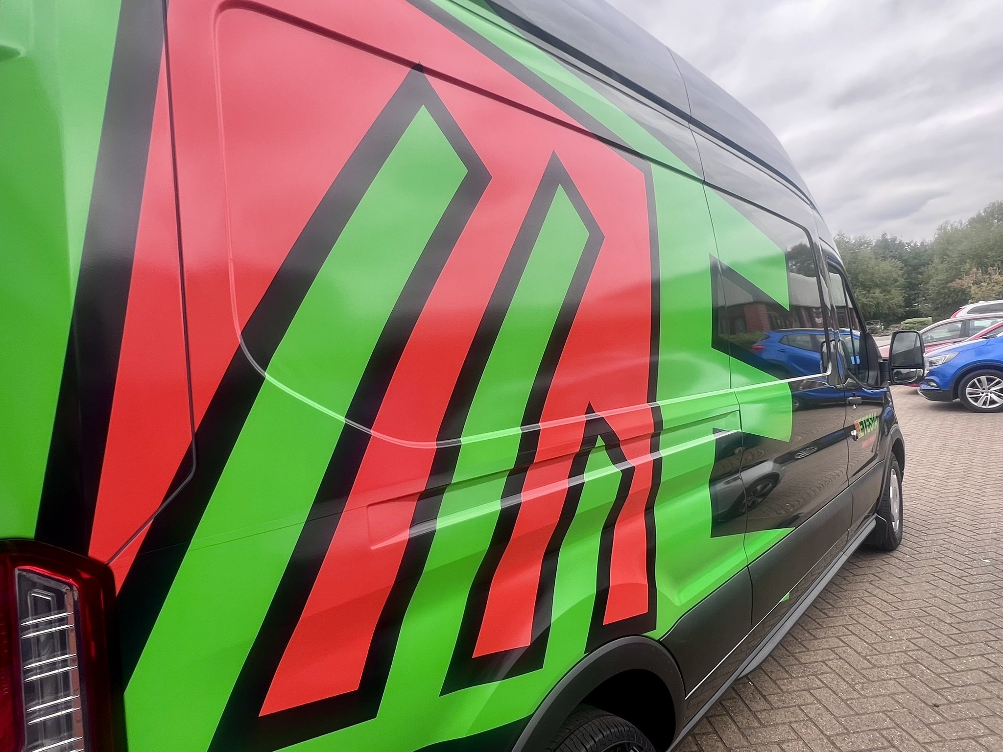 A black van with green and red decorative graphics on its side, parked on a brick pavement in a lot with other vehicles and trees in the background.