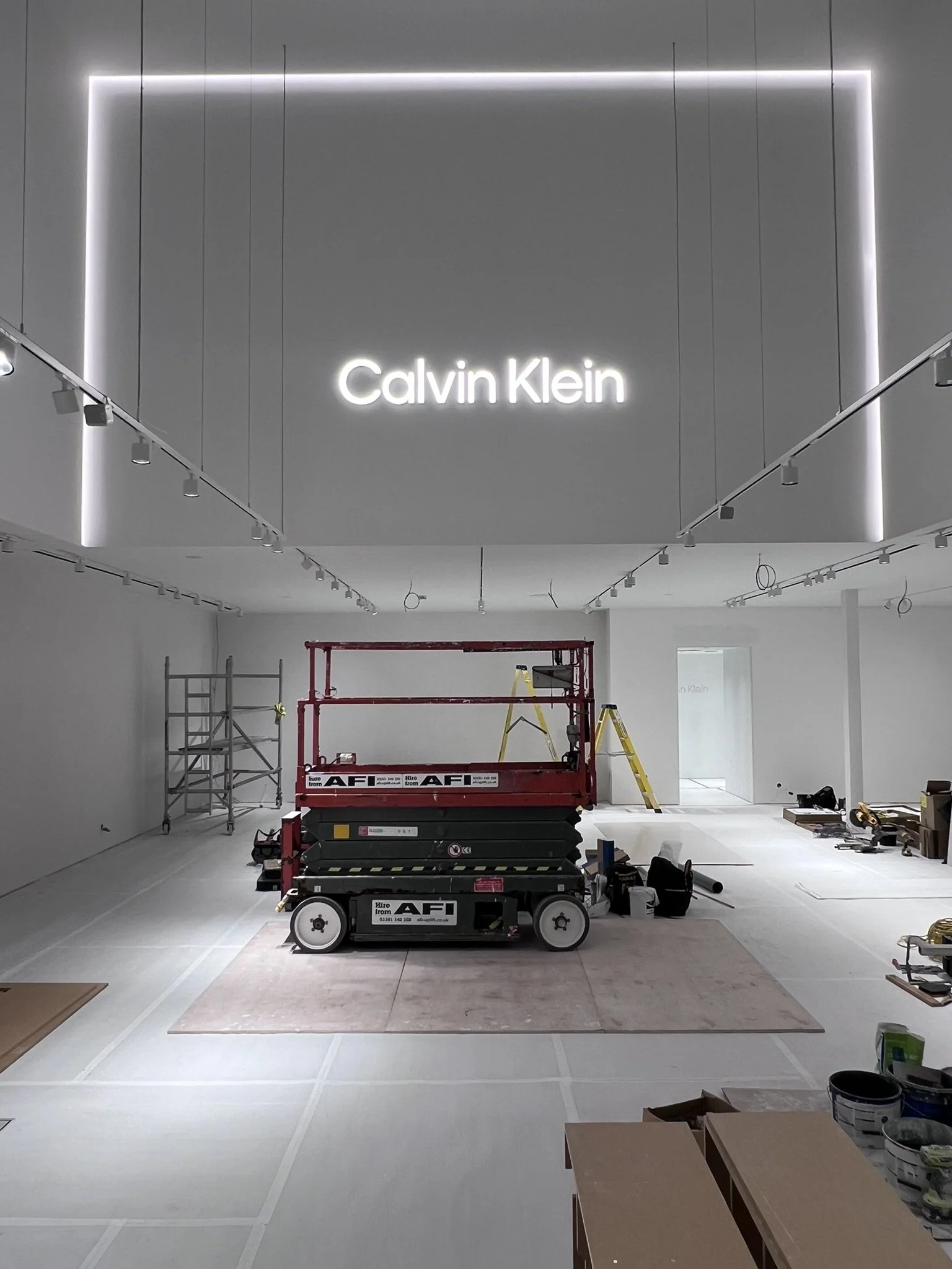 Interior of a retail store under construction, with scaffoldings, tools, and a lift machine in the center, and a prominent Calvin Klein sign illuminated on the wall.