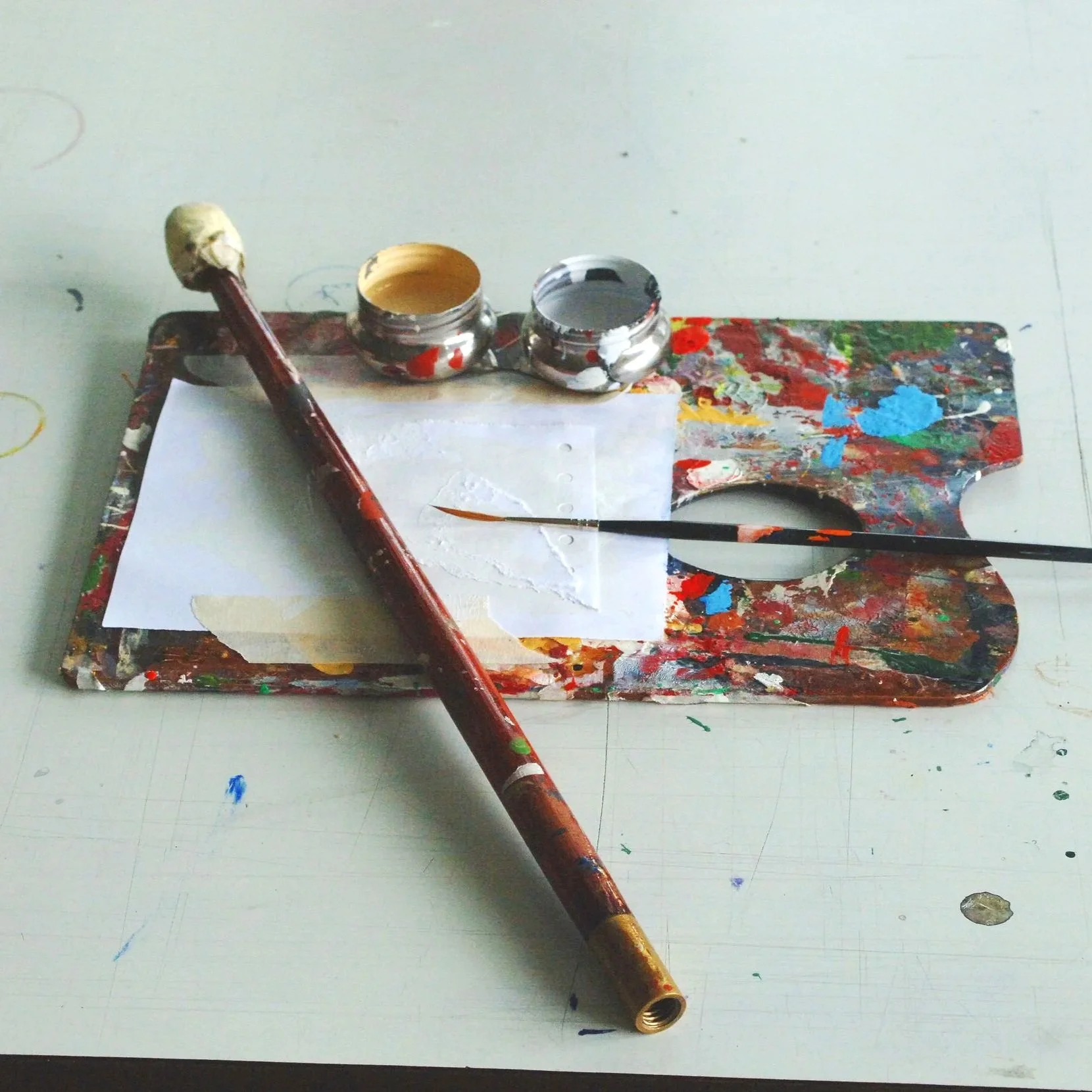 Paint palette with splatters, a paintbrush, and small open containers of beige and black paint on a white surface.