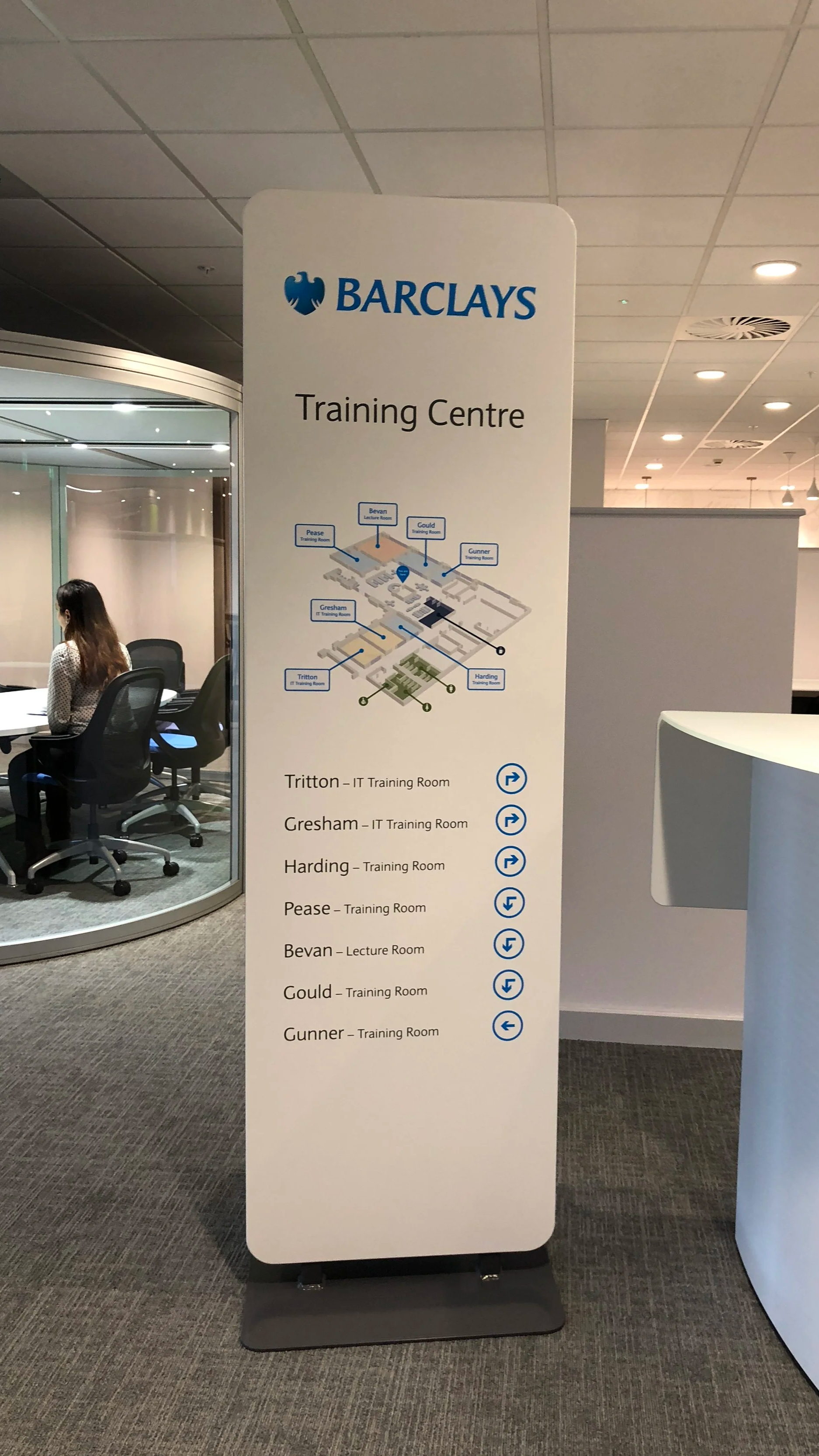 A tall freestanding sign listing training rooms at Barclays with a map and directional arrows in a corporate office setting.