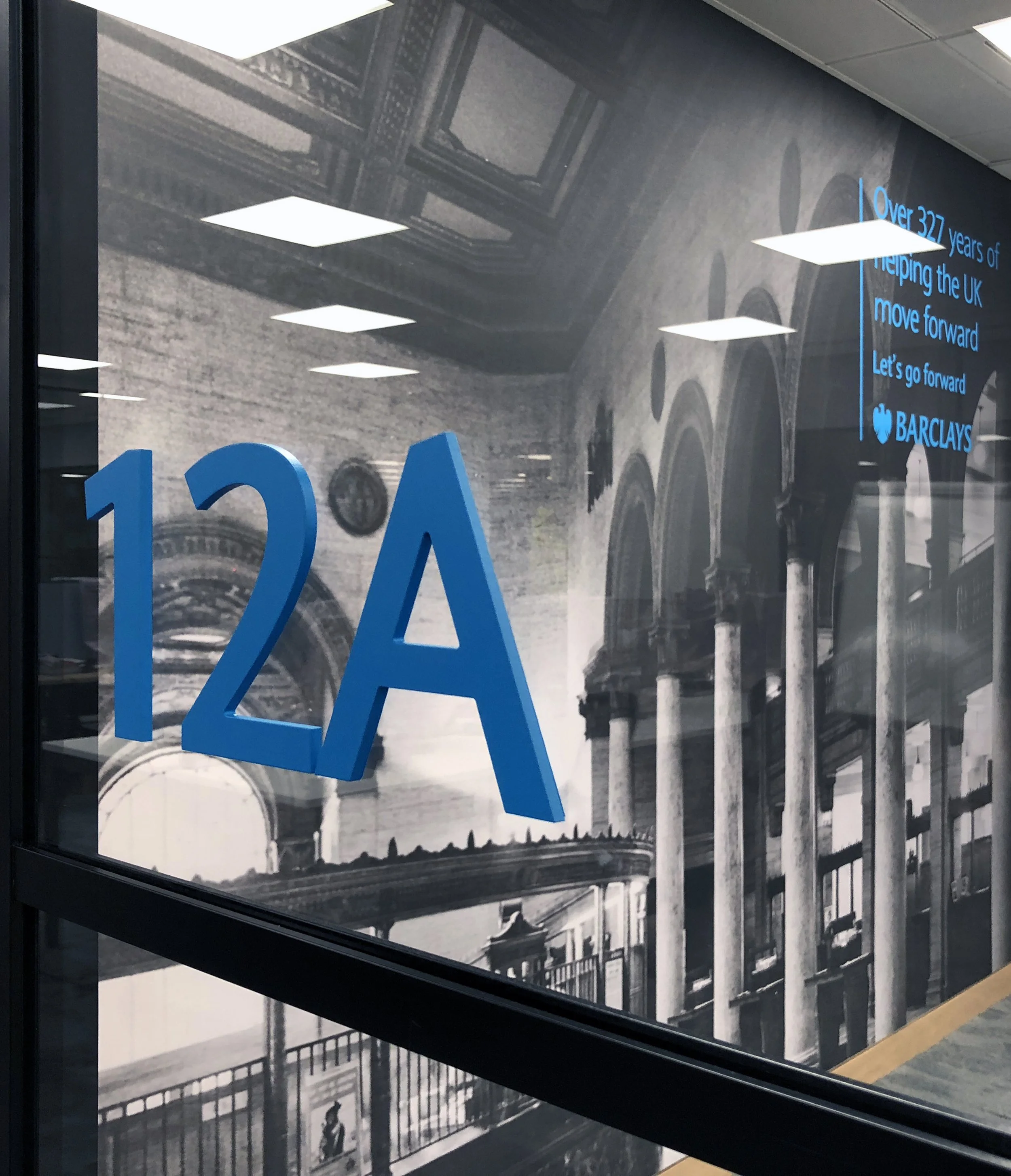 View of an office window with large blue text reading '12A'. Behind the window, there is a black and white mural of classical architecture, including arches, columns, and detailed ceiling designs. In the upper right corner, there is a blue text overlay that says 'Over 327 years of helping the UK move forward. Let's go forward. Barclays'.