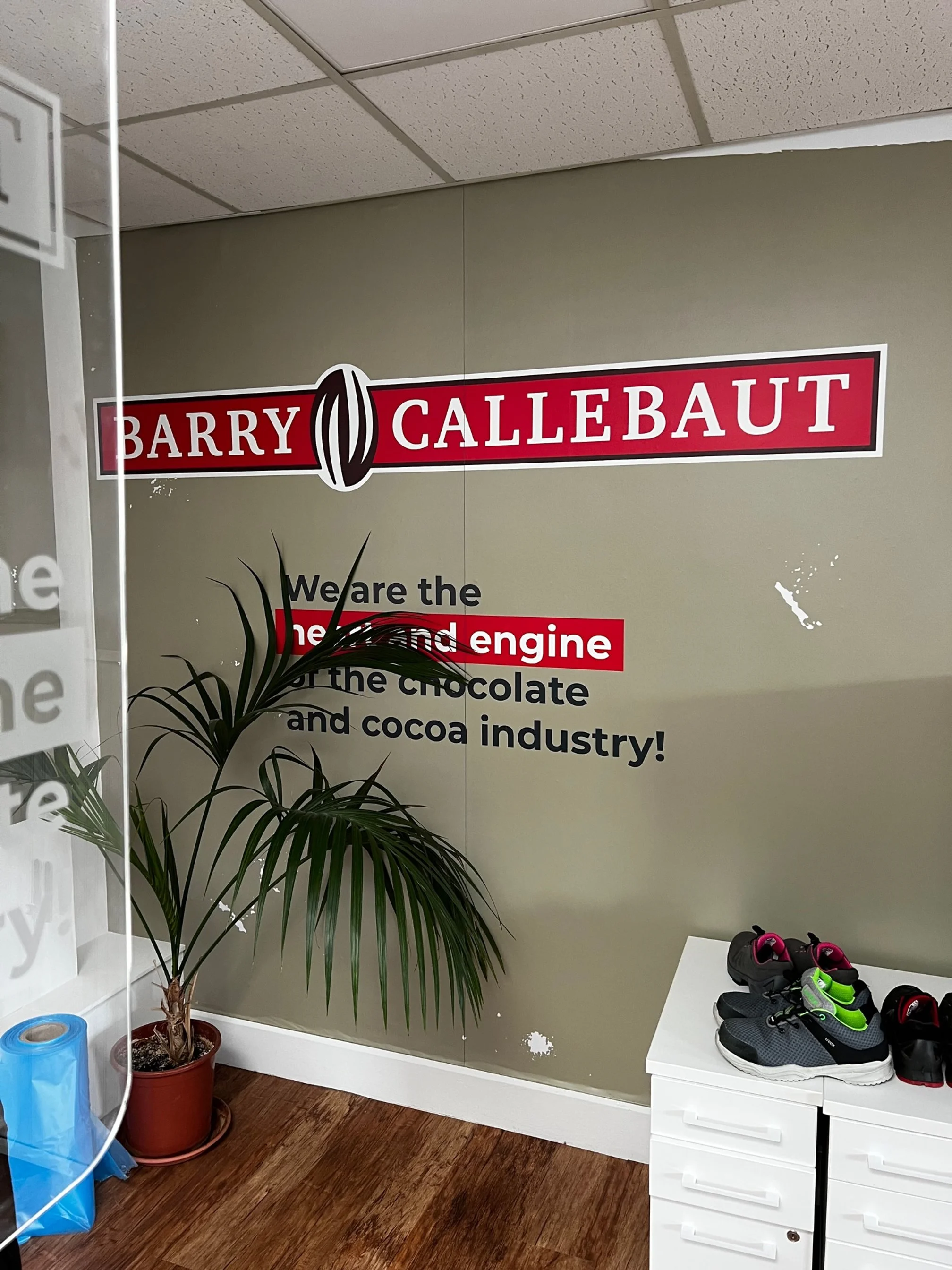 Sign on wall with the name Barry Callebaut, a chocolate and cocoa industry company, and a message stating 'We are the heart and engine of the chocolate and cocoa industry!' includes a potted plant, shoes, and a blue plastic bag in an indoor setting.