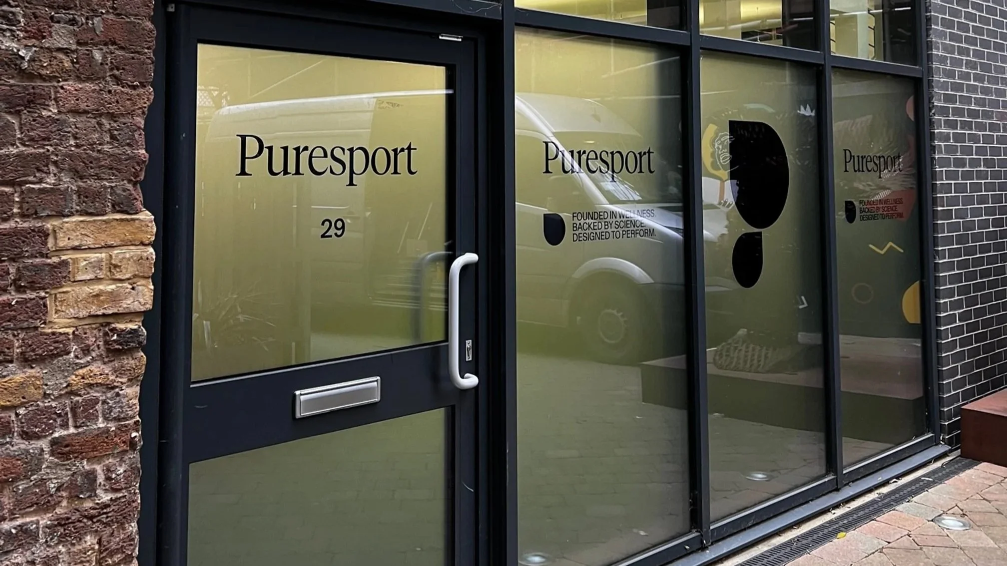 Storefront with glass doors and windows labeled 'Puresport' in black text. Reflection of a white van and street scene visible on the glass. The storefront has a brick wall on the left and black framing.