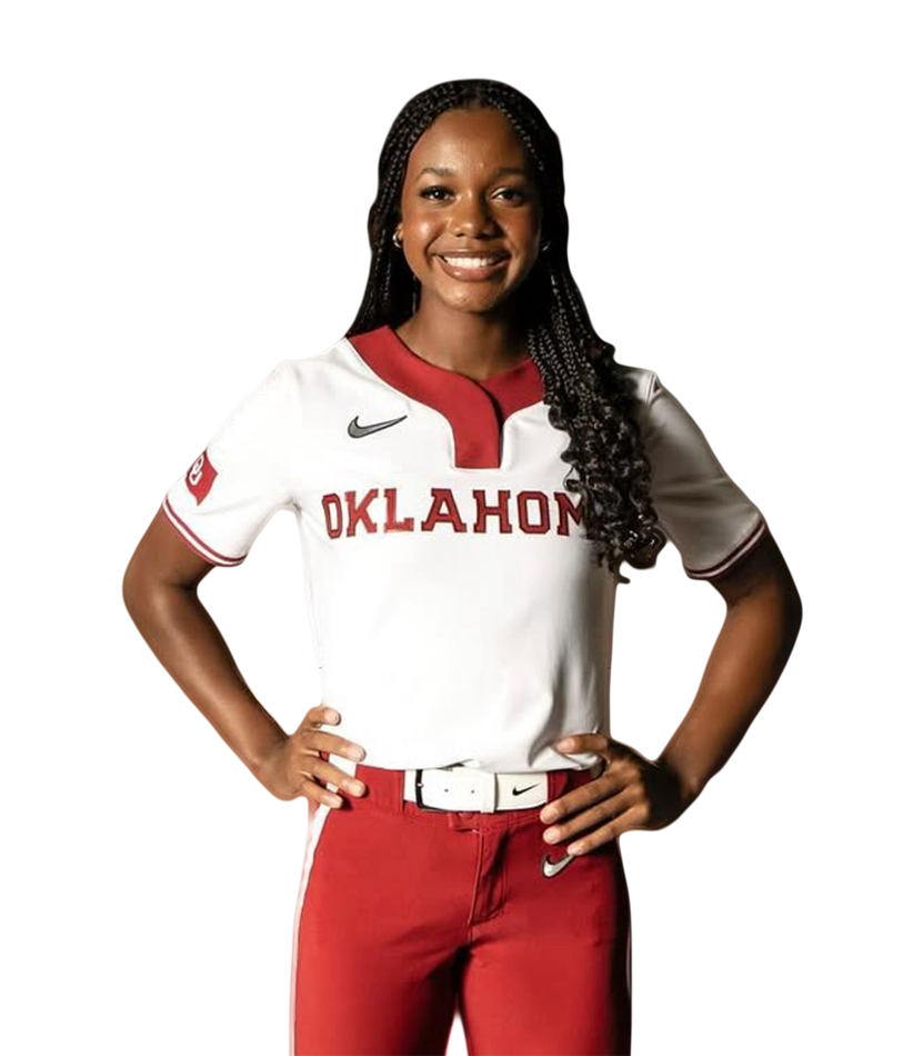 A young woman wearing a white Oklahoma softball jersey with red accents, red pants, and a white belt, standing with her hands on her hips and smiling.