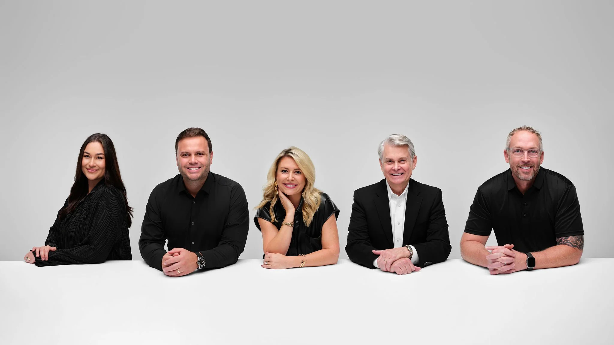 Group of five diverse people in business casual and formal attire, sitting at a white table against a plain gray background, smiling and looking at the camera.