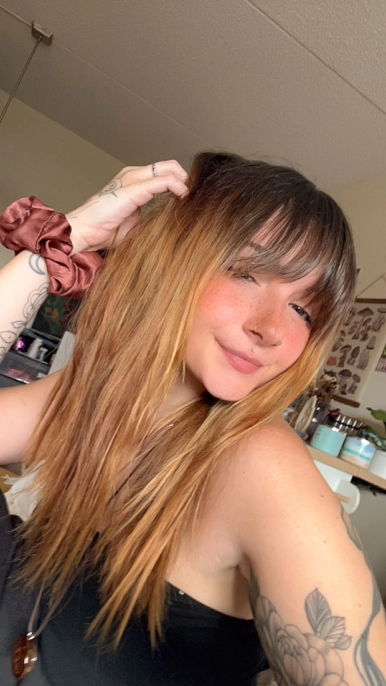 A woman with long, light brown hair, freckles, a septum piercing, and tattoos on her arm, smiling and winking, in a room with various decorations and supplies in the background.