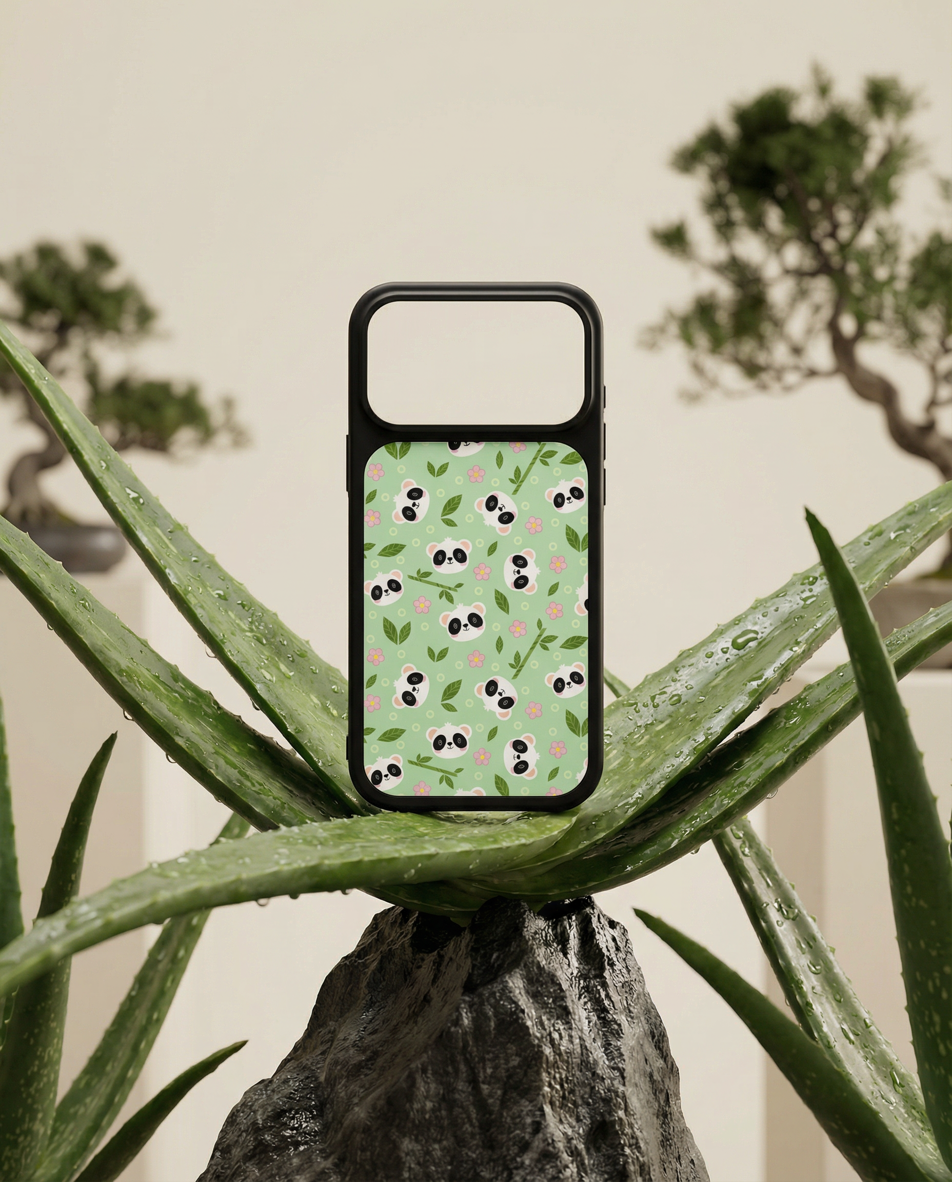 A phone case with panda and pink flower patterns placed on a green aloe vera plant amidst other potted plants in the background.