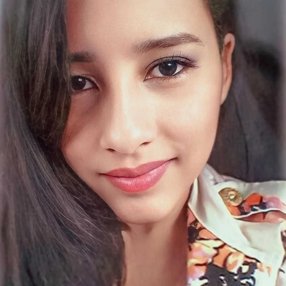 Close-up of a woman with long dark hair, blue eyes, and pink lipstick, wearing a floral shirt.