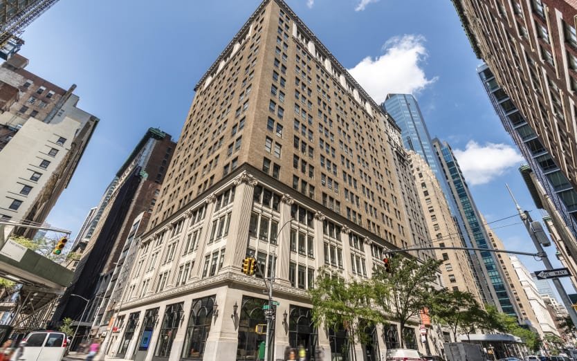 A city street view with tall buildings, including a prominent brown and white building, and a blue sky with some clouds.
