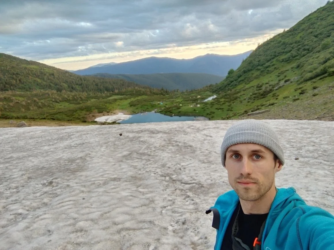A man wearing a gray beanie and blue jacket taking a selfie on a snowy mountainside with a green valley, small lakes, and mountains in the background.