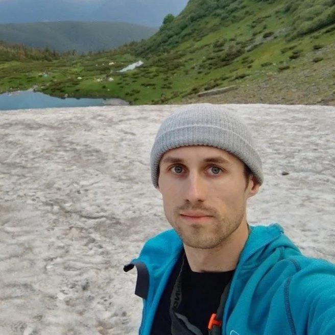 A man taking a selfie on a snowy landscape with green hills and lakes in the background.