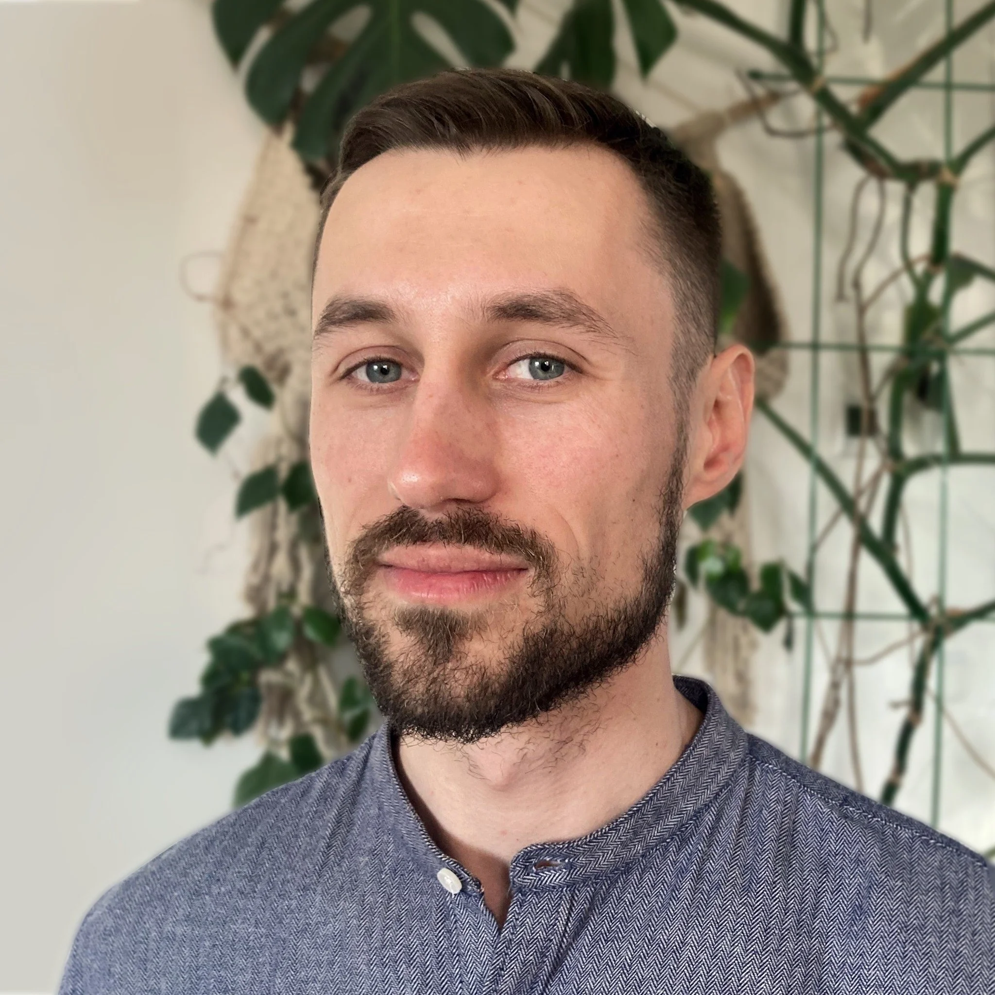 A young man with short brown hair, blue eyes, and a beard, wearing a gray button-up shirt, standing indoors with green leafy plants in the background.