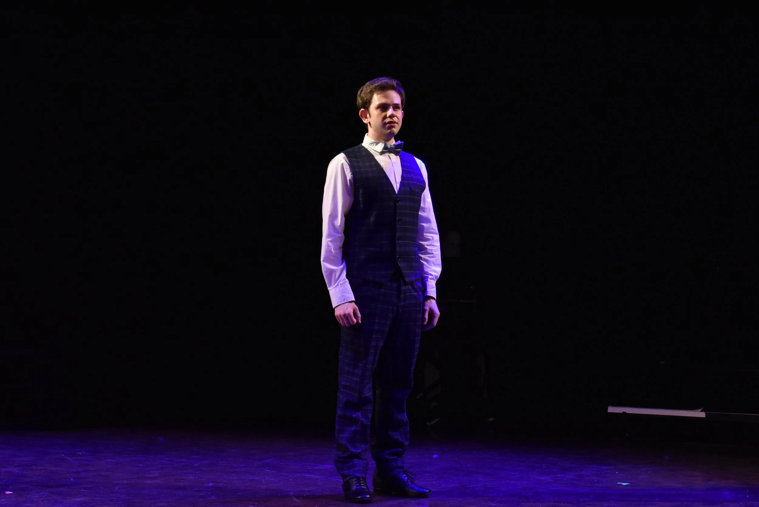A young man standing on a dark stage, dressed in a suit with a bow tie, illuminated by stage lighting.