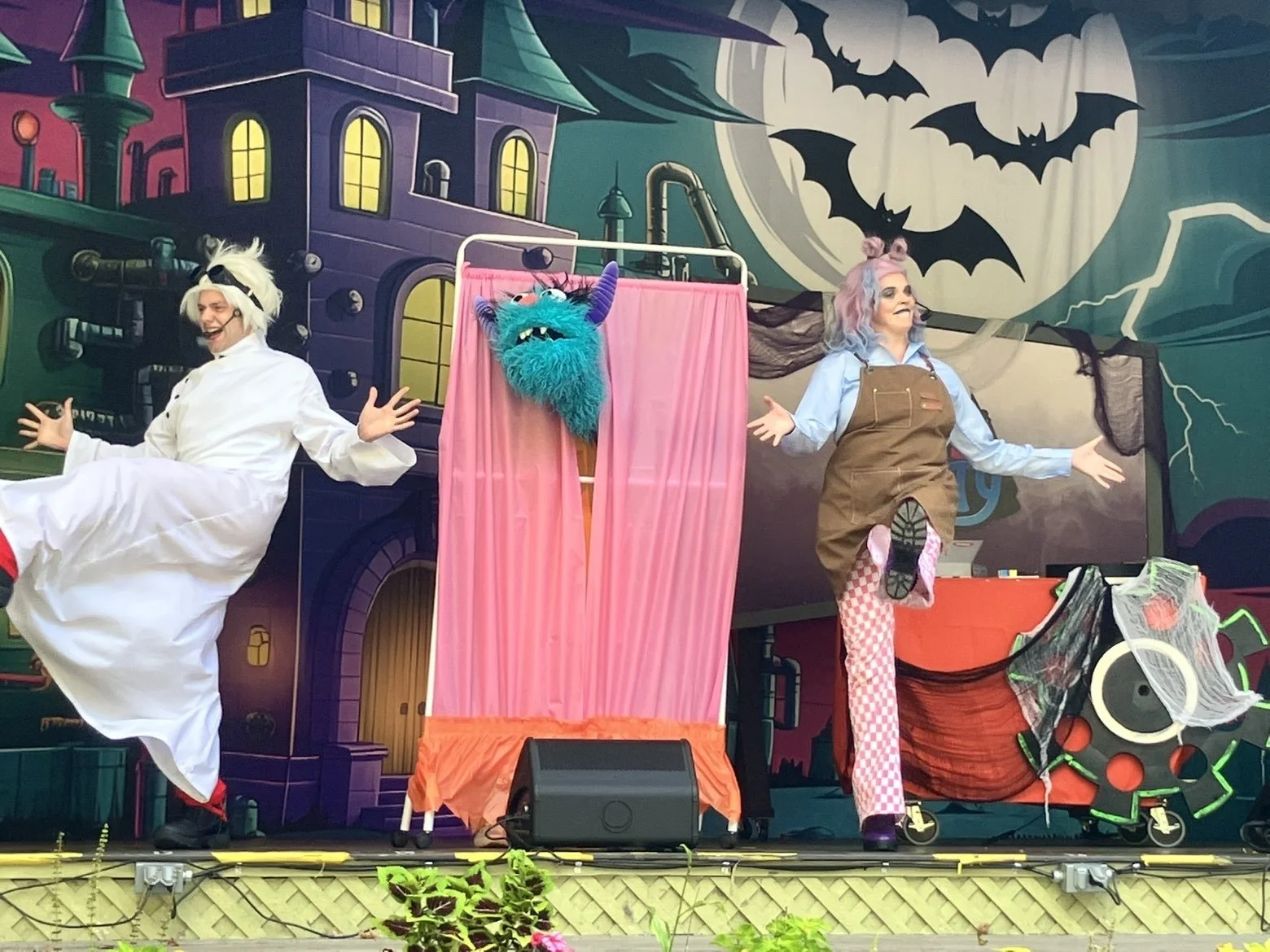 Three performers on stage dressed in colorful costumes with a Halloween-themed backdrop featuring a full moon and flying bats. The stage has a puppet with a blue furry face, purple horns, and a big smile, framed by pink curtains. The performers are animated, smiling, and engaging with the audience.