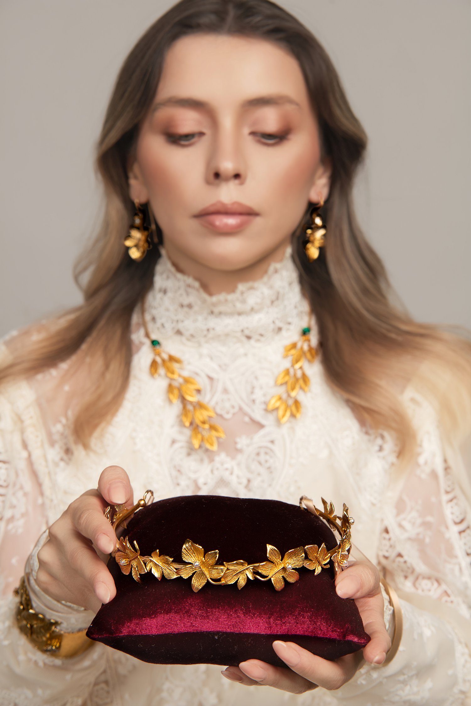 A woman wearing a lace dress and gold jewelry, holding a velvet pillow with a gold floral tiara.