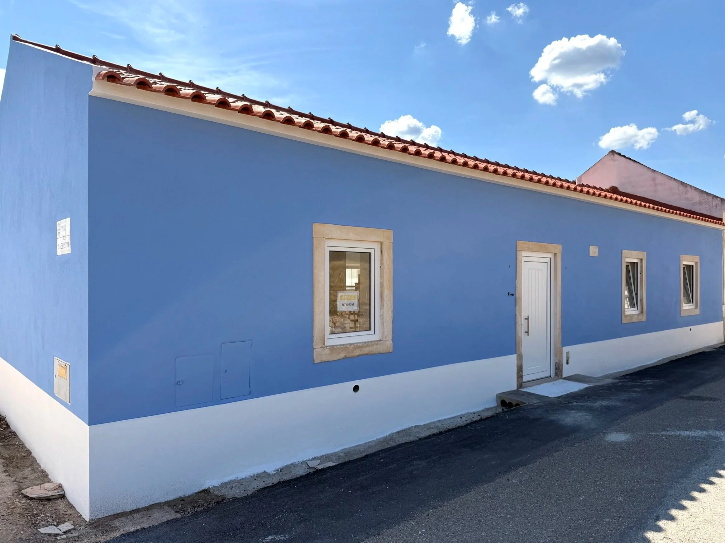 Casa de parede azul com telhado de telhas vermelhas, uma porta branca ao centro, três janelas com moldura de pedra e céu azul com nuvens.