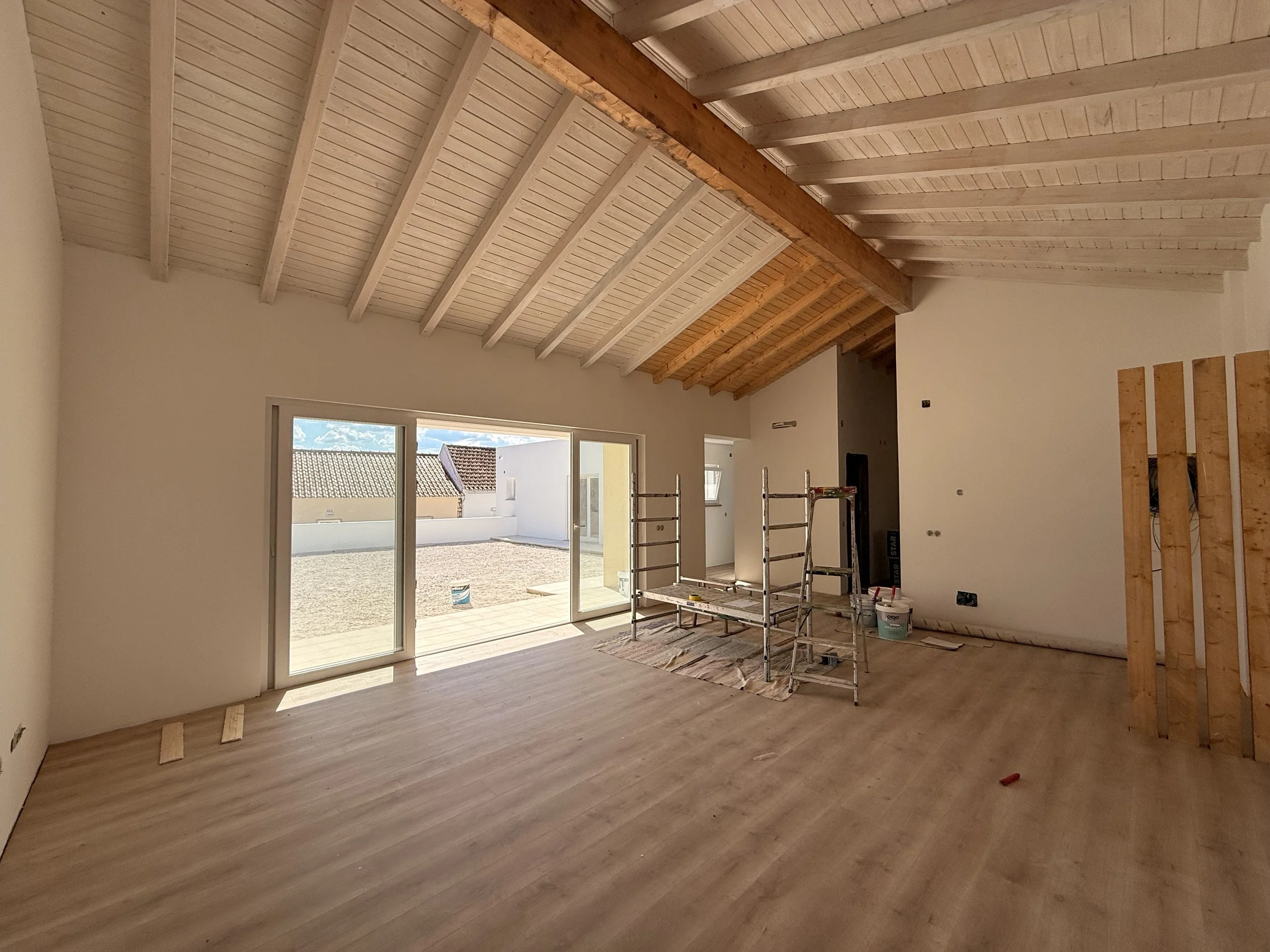 Sala de estar em construção com teto de madeira, piso de assoalho de madeira, grande porta de vidro que dá para uma área externa, alguns materiais de construção e escada de andaime no centro.