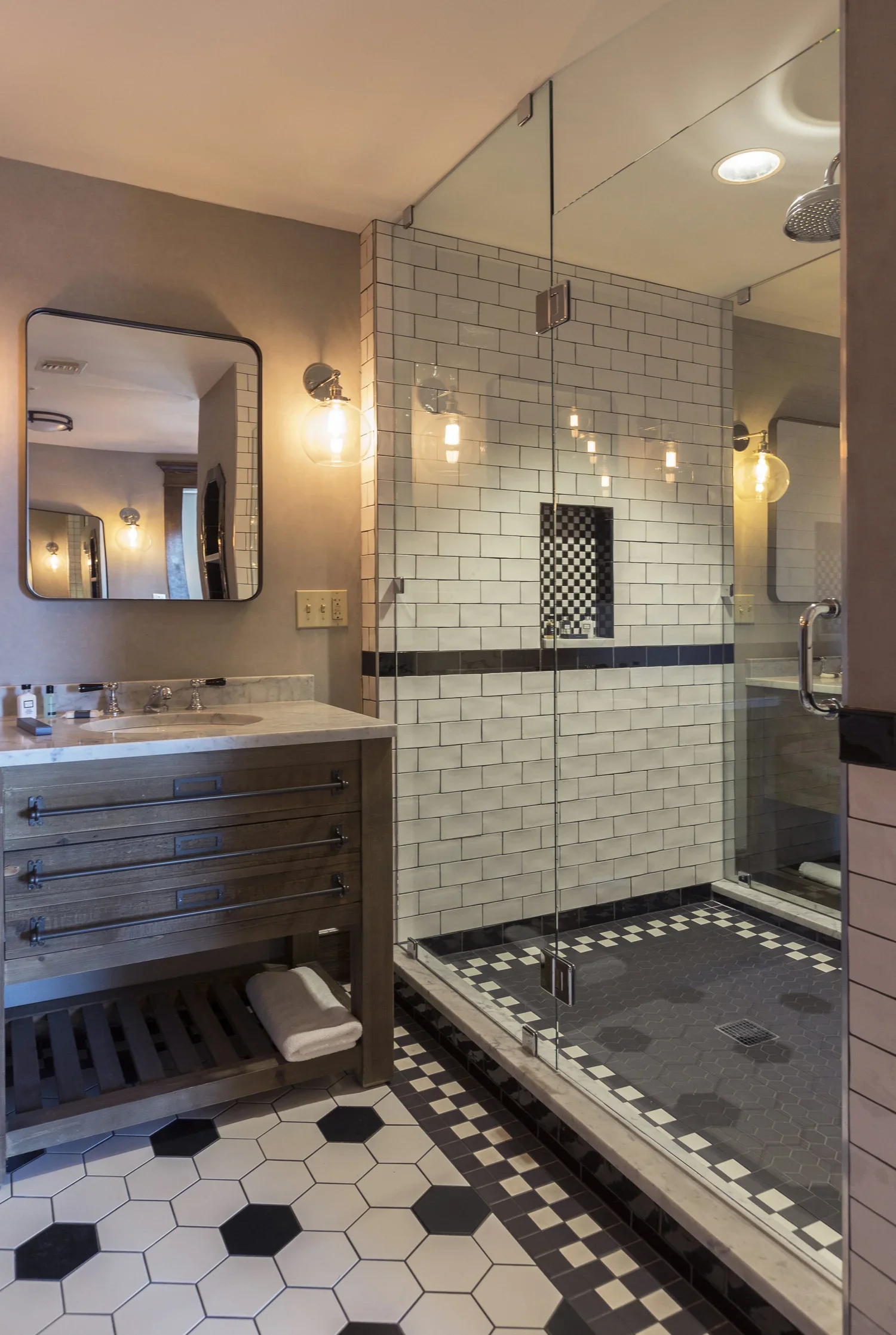 Modern bathroom with a glass shower enclosure, white subway tile walls, a wooden vanity with a marble countertop, a mirror, and black and white hexagonal tile flooring.