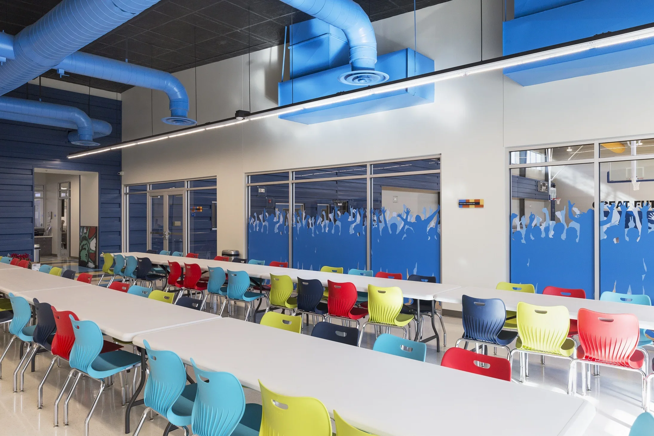 Colorful classroom or cafeteria with multicolored chairs around white tables, large windows with blue decorative graphics of cheering crowd, blue industrial ductwork on ceiling, modern interior design
