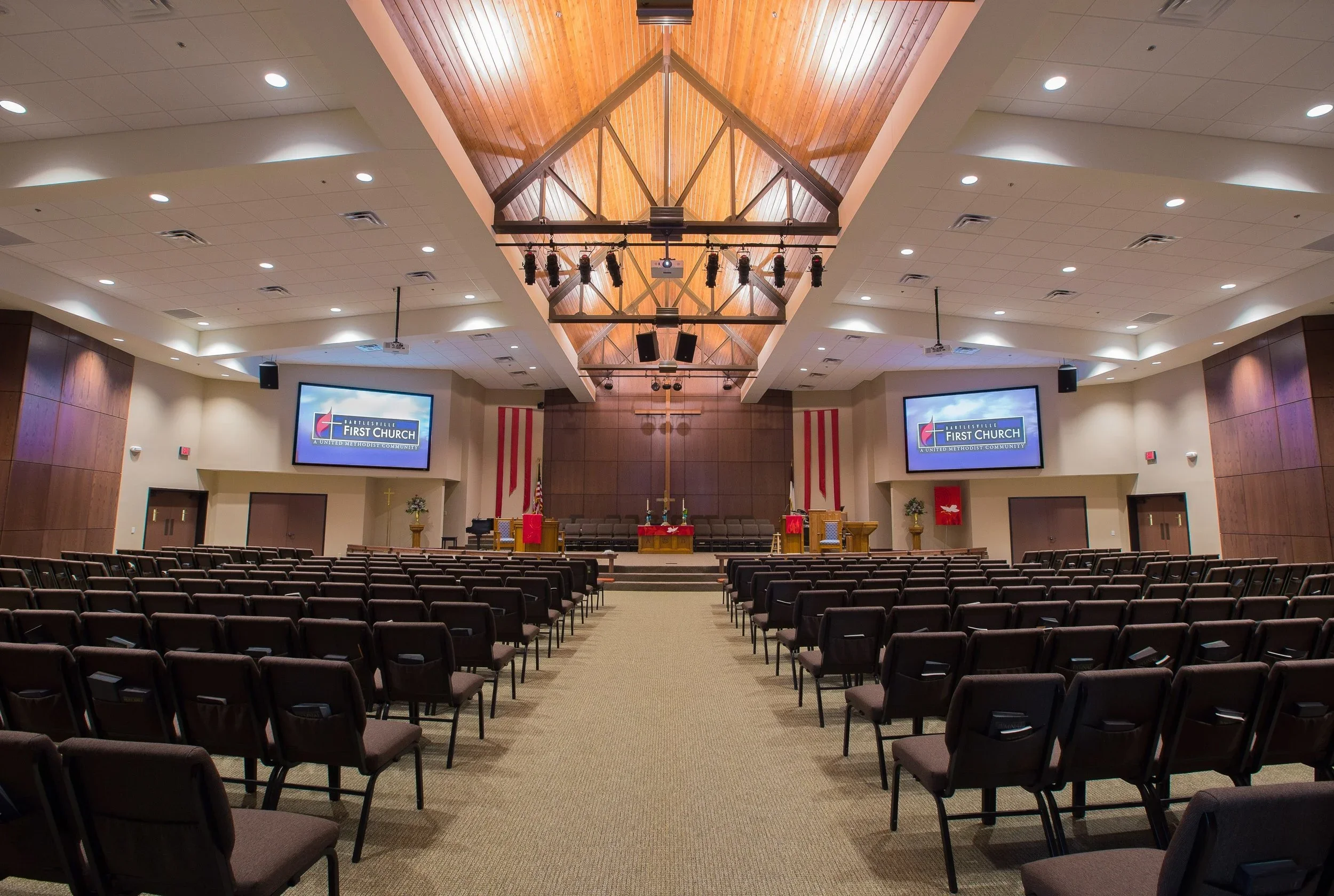 Empty church auditorium with rows of brown chairs facing an elevated stage with a cross, flowers, and American flags. Two large screens display 'First Church' logos.