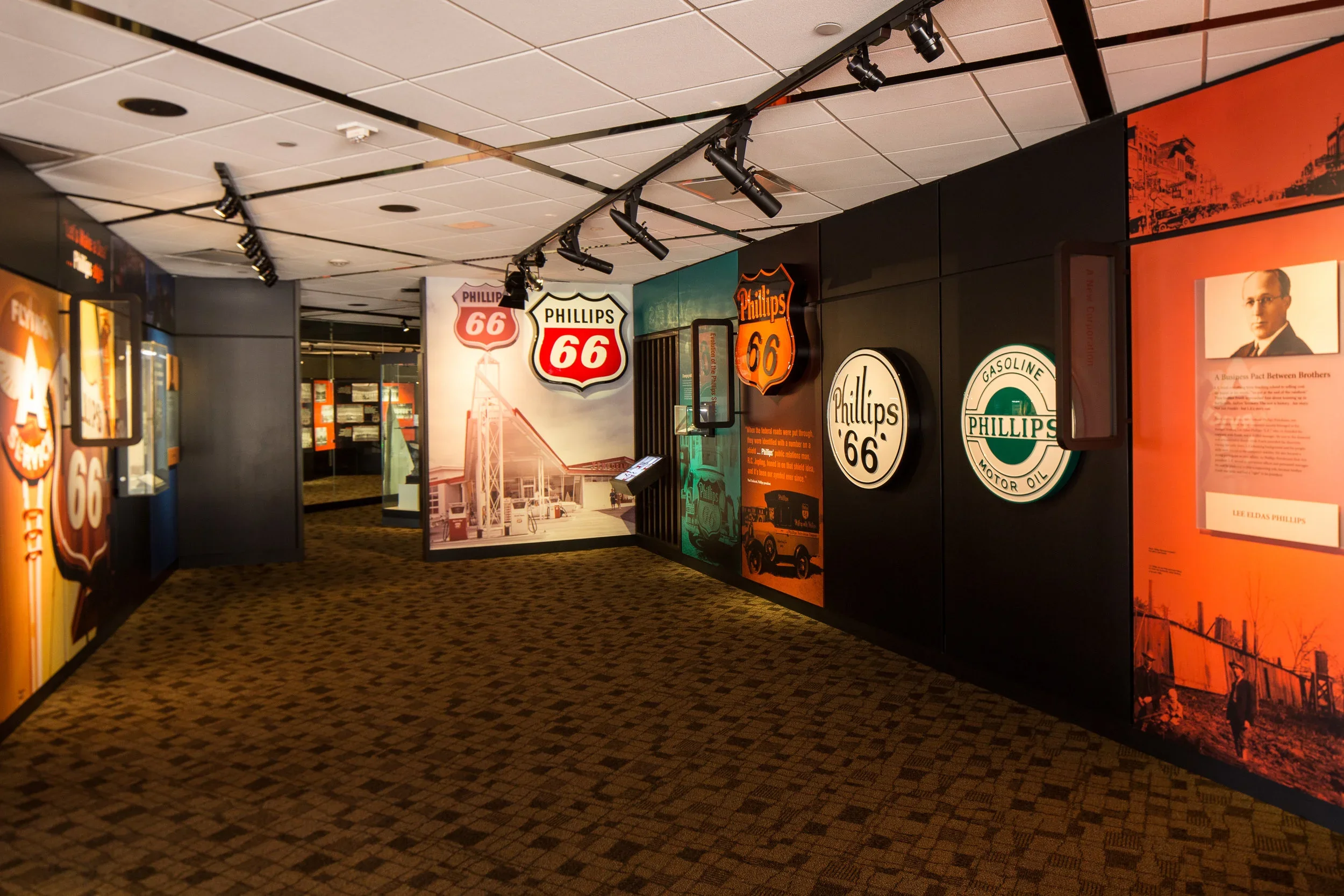 Museum exhibit featuring vintage signs related to Phillips 66 gas station, including signs with logos, historical photos, and informational displays, in a room with dark walls, track lighting, and patterned carpet.