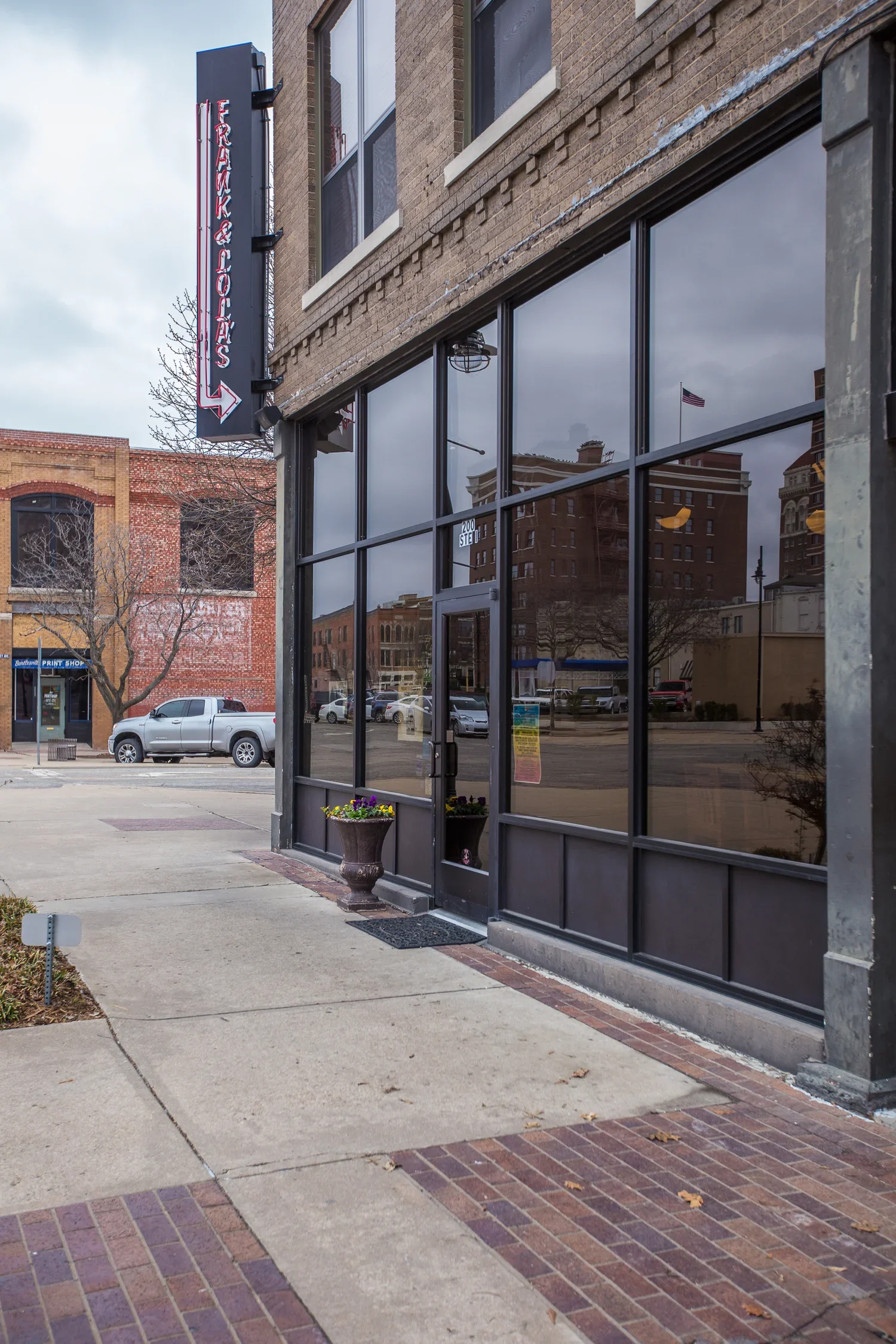 The exterior of a building with a large glass storefront and a vertical sign with an arrow pointing left, alongside a sidewalk and parked cars in the background.