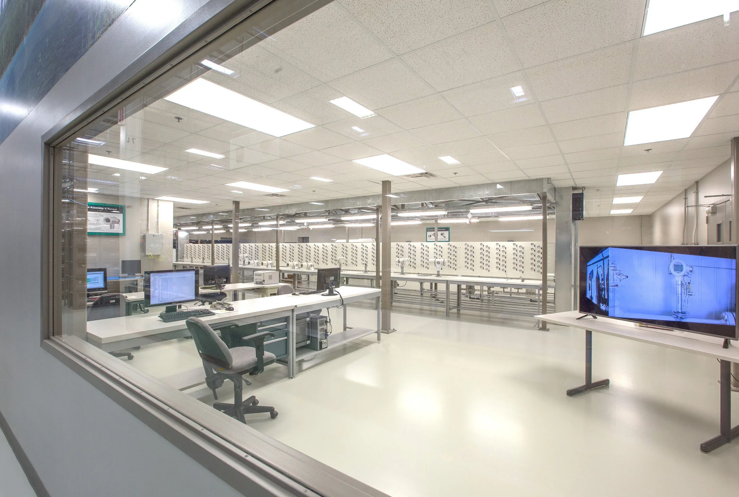 View of a robotics laboratory through a large window. Inside, there are workstations with computers, chairs, and technical equipment. In the background, there are numerous robotic arms or devices arranged in rows, and a large screen on a stand displa