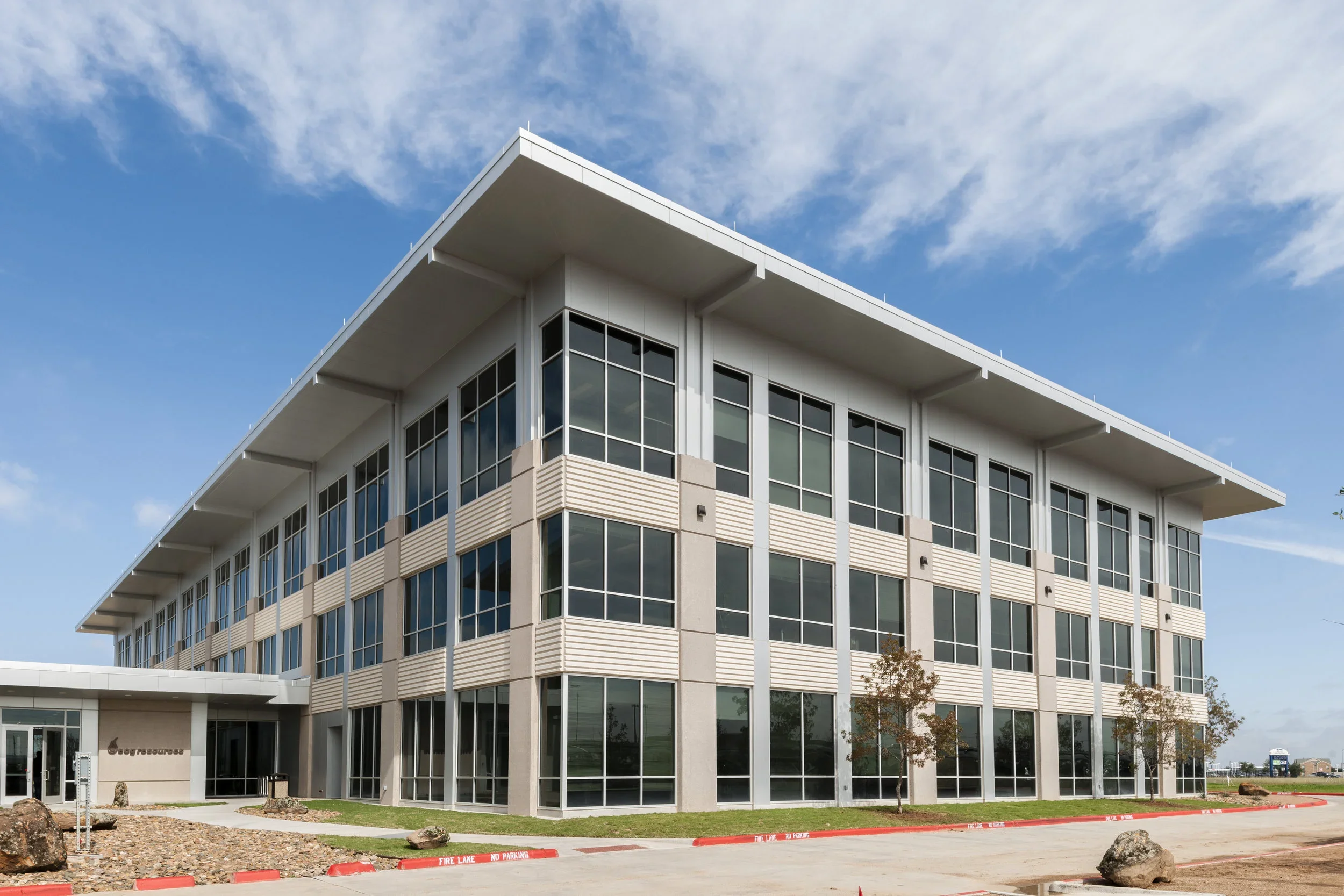Modern three-story office building with large glass windows, beige accents, and a flat roof, under a partly cloudy blue sky.