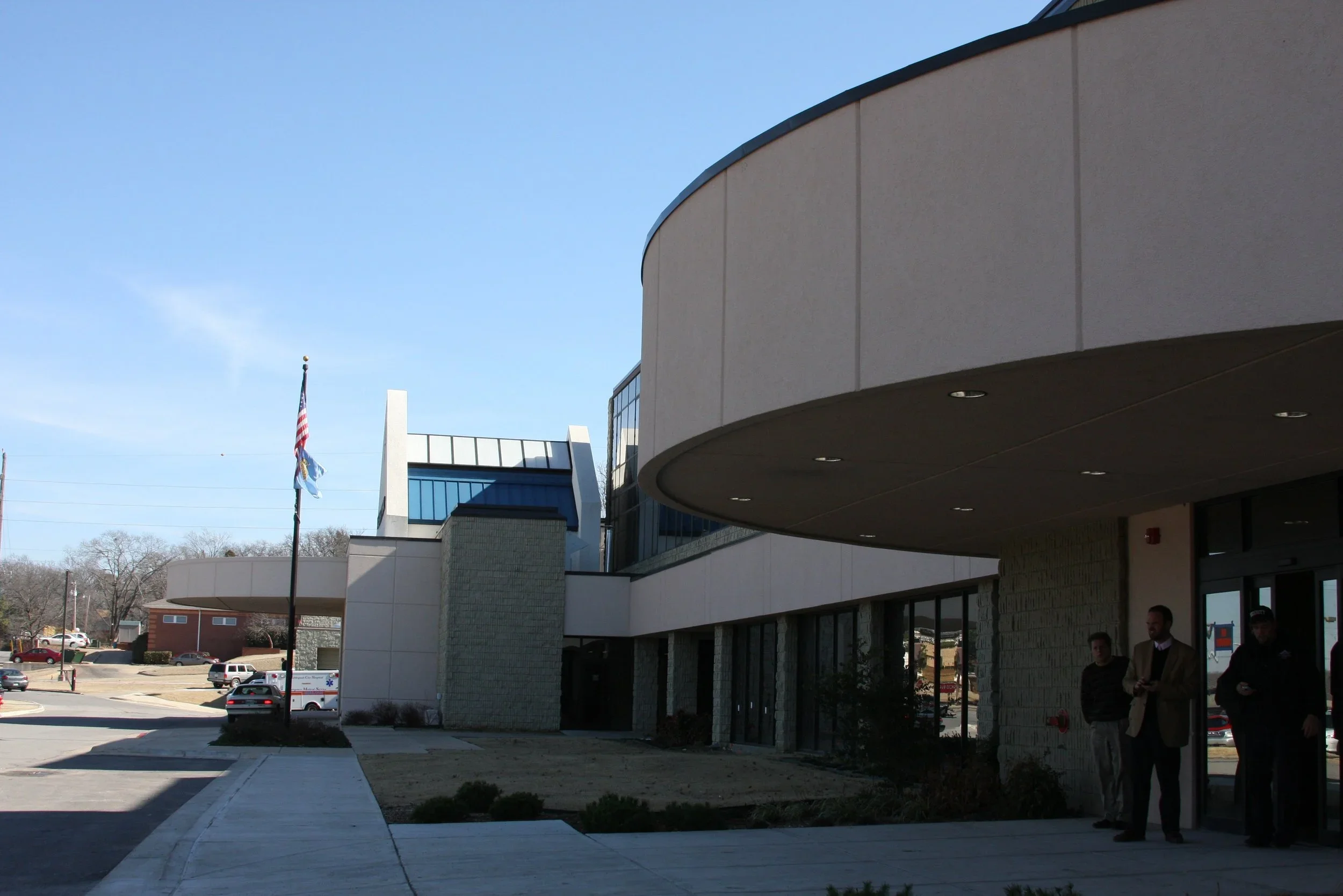 Modern commercial building with large glass windows, concrete and brick exterior, and a curved overhang. There are three men standing near the entrance, and a flagpole with a flag in the foreground.