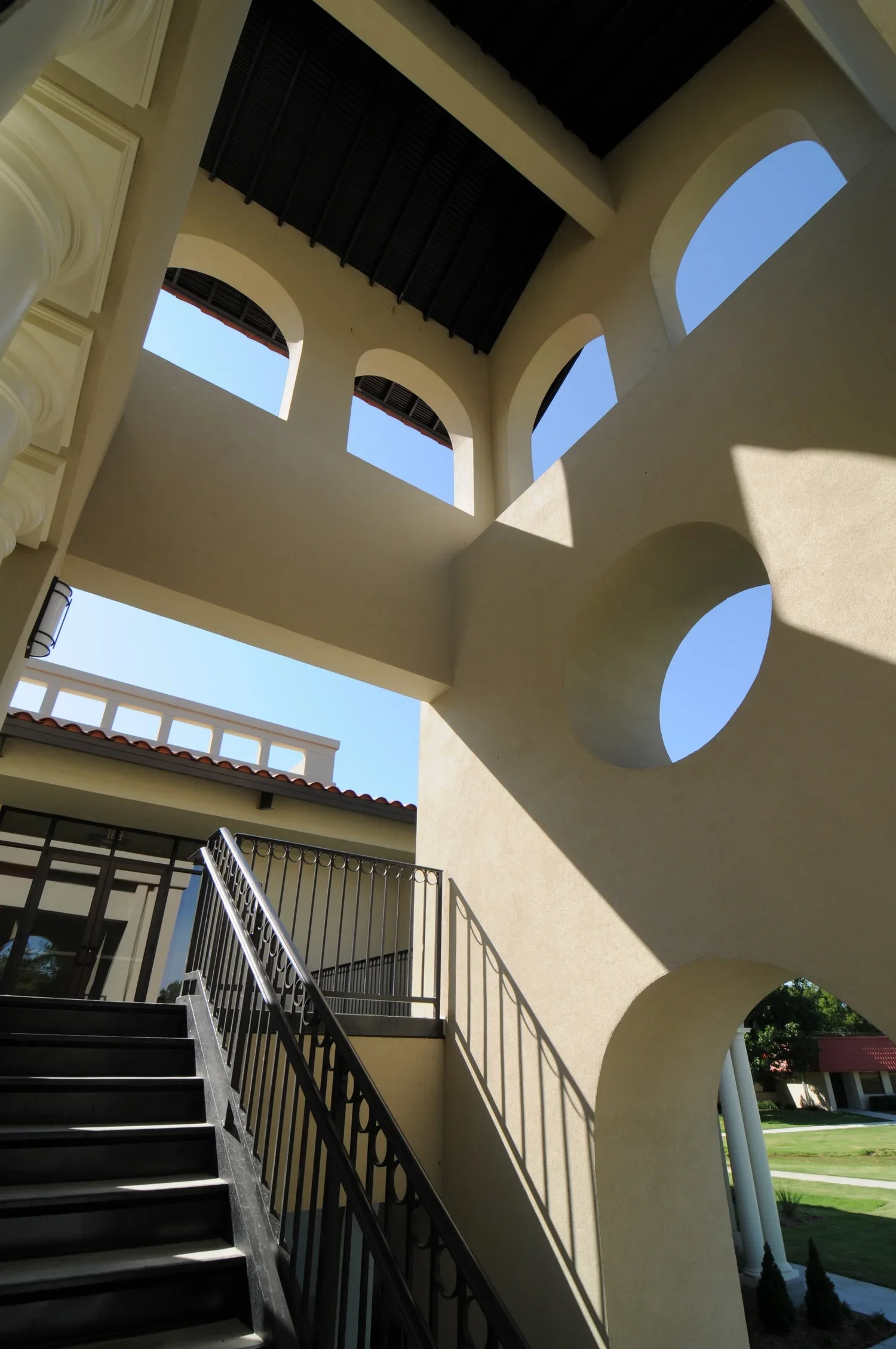 Photo of a building's exterior featuring arched windows and circular architectural cutouts, with a staircase leading up to a balcony and a view of a grassy lawn and other buildings in the background.