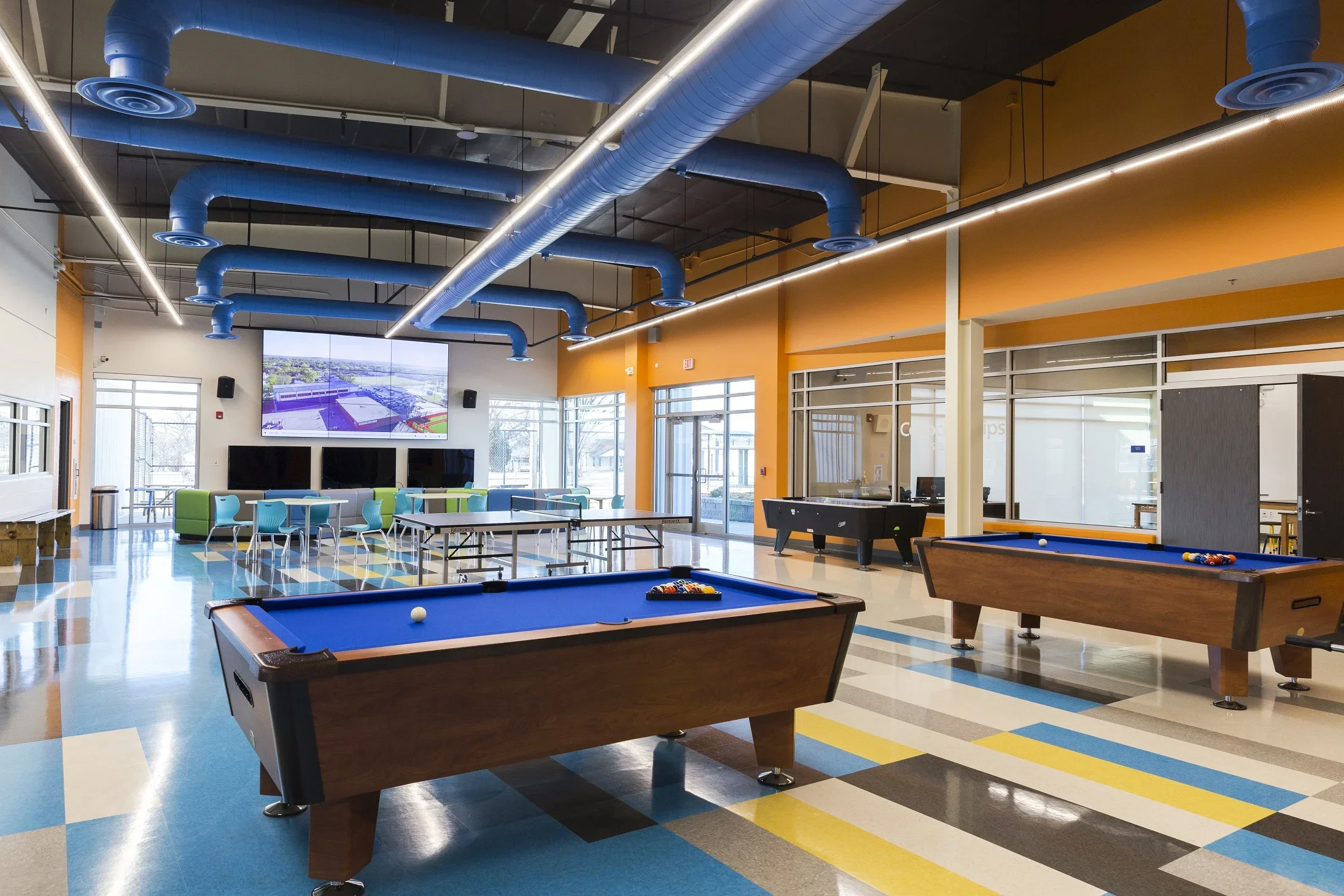 Interior of a recreational room with two pool tables, seating area, large TV, foosball table, and colorful floor and wall decor.