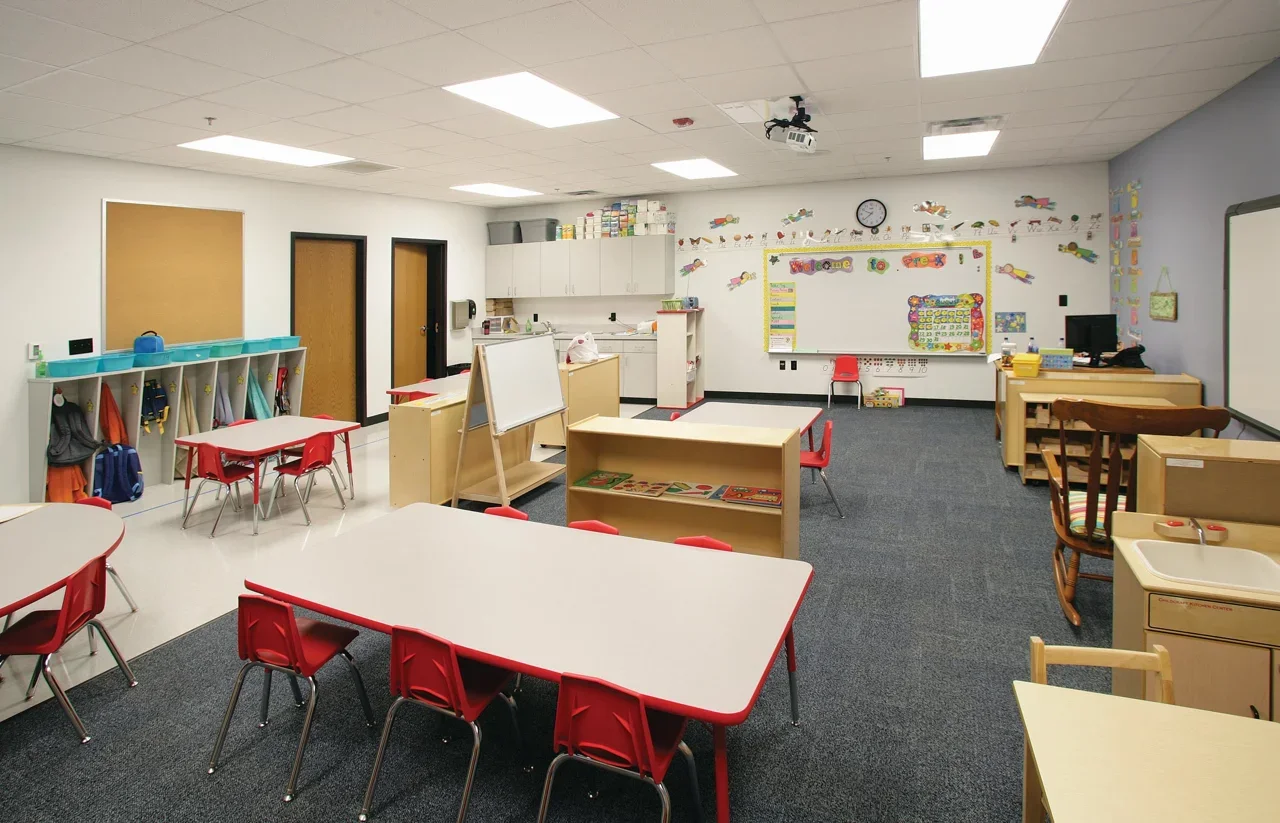 Empty preschool classroom with tables, chairs, educational materials, and colorful wall decorations.