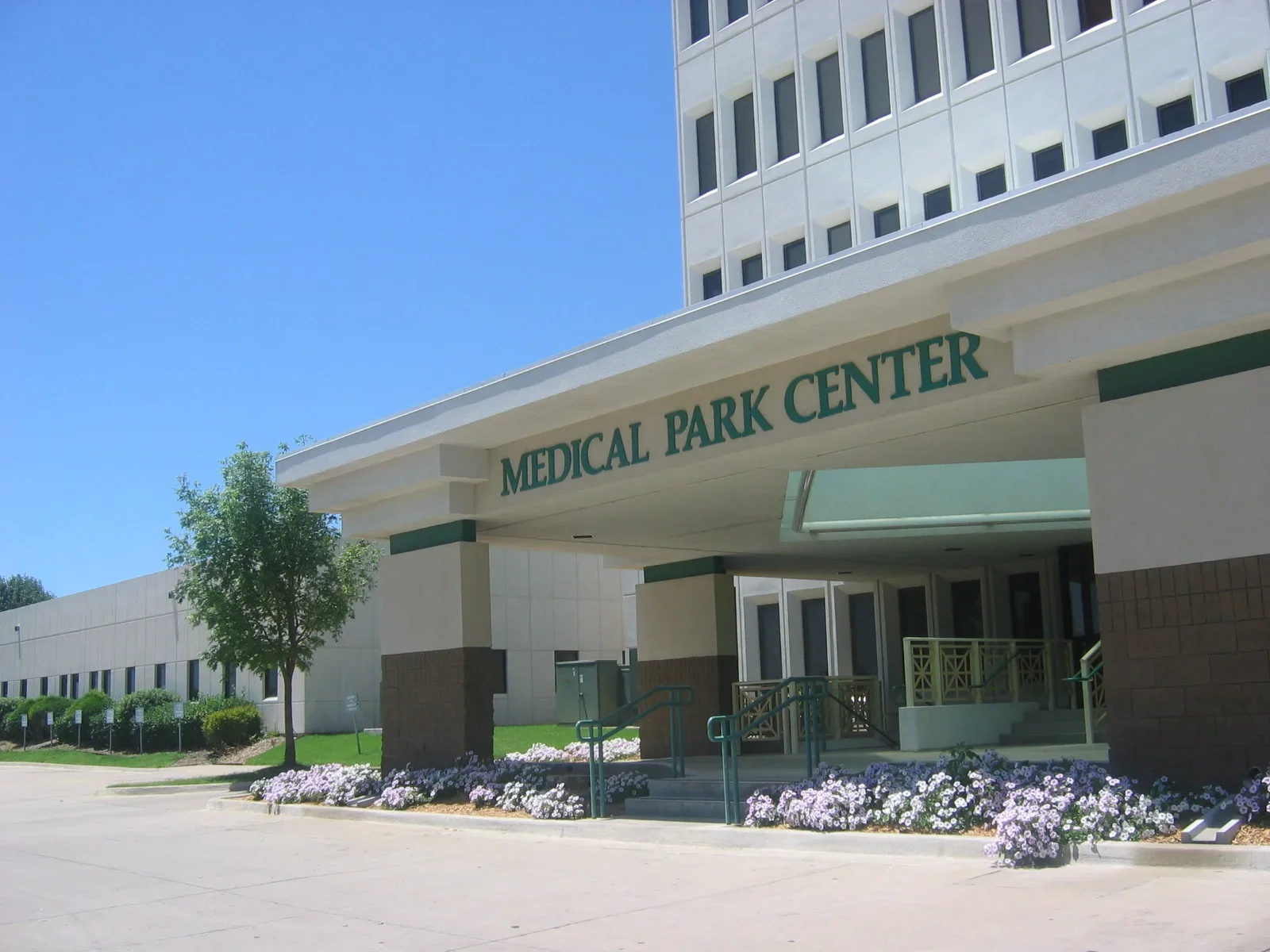 Outside view of Medical Park Center building with flowers and trees in front, clear blue sky.
