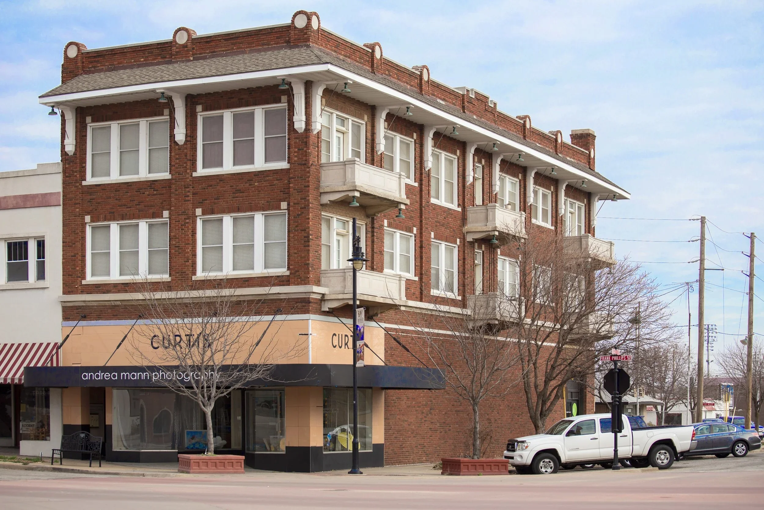 A three-story brick building with a corner storefront on a city street, with leafless trees, parked cars, and utility poles, under a partly cloudy sky.