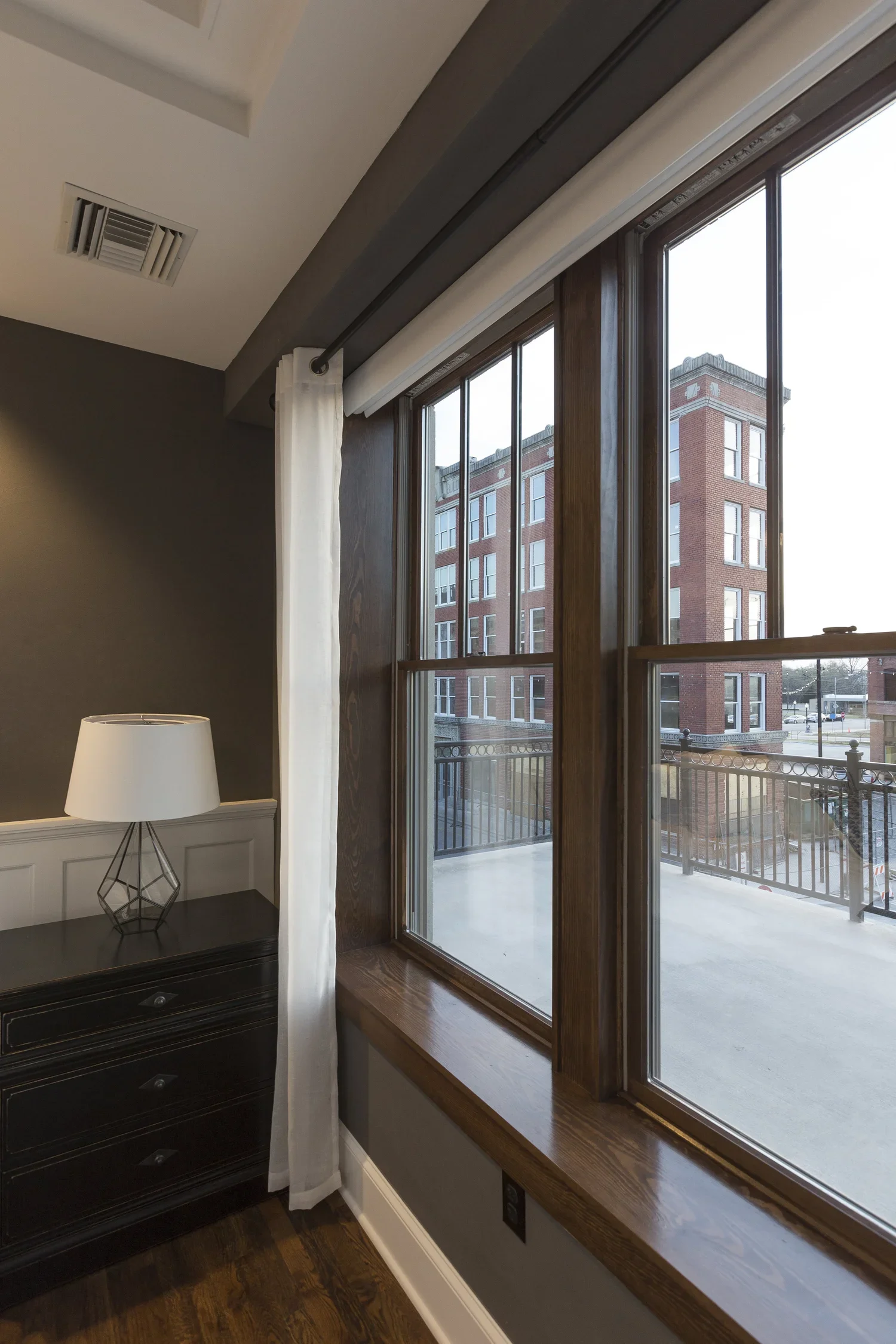 Interior view of a room with a large wooden window, a white curtain, a black dresser with a lamp, and a view of a brick building outside.