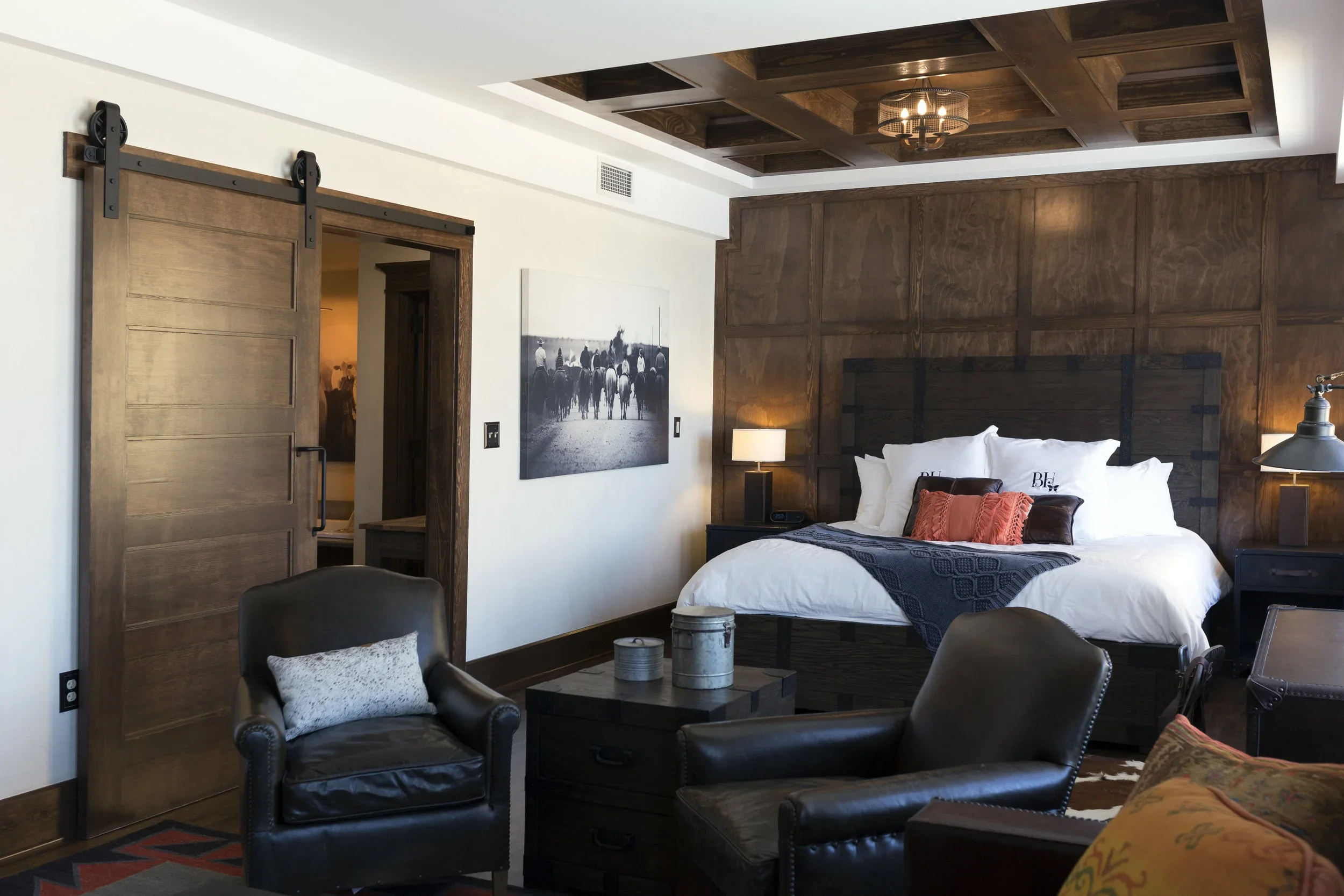 A cozy bedroom with a large bed, dark wood headboard, and white bedding. Two black leather armchairs, a small table with decorative items, and a white and black artwork on the wall. Wooden sliding barn door and wood-paneled ceiling. Warm lighting fro