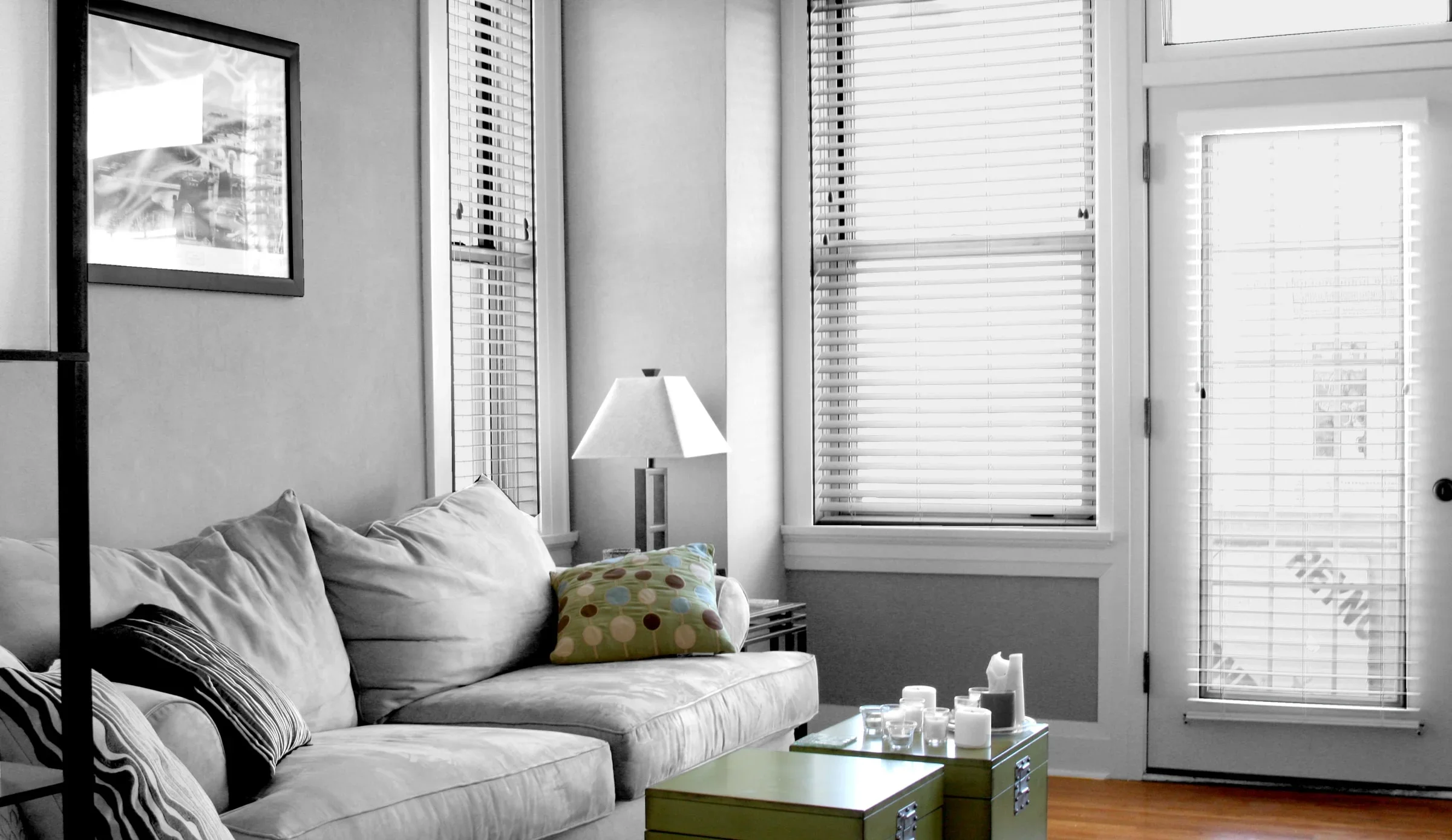 Living room with a beige sofa, colorful pillow, side table with candles, lamp, and windows with blinds, black and white photograph on the wall, and door with blinds.