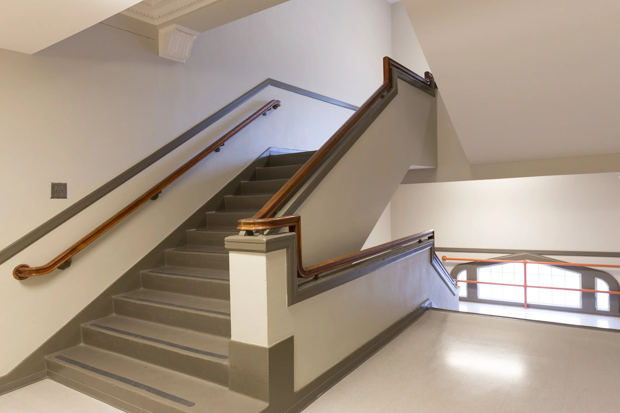 Indoor staircase with gray steps, wooden handrails, and beige walls, leading to an upper and lower floor with windows.