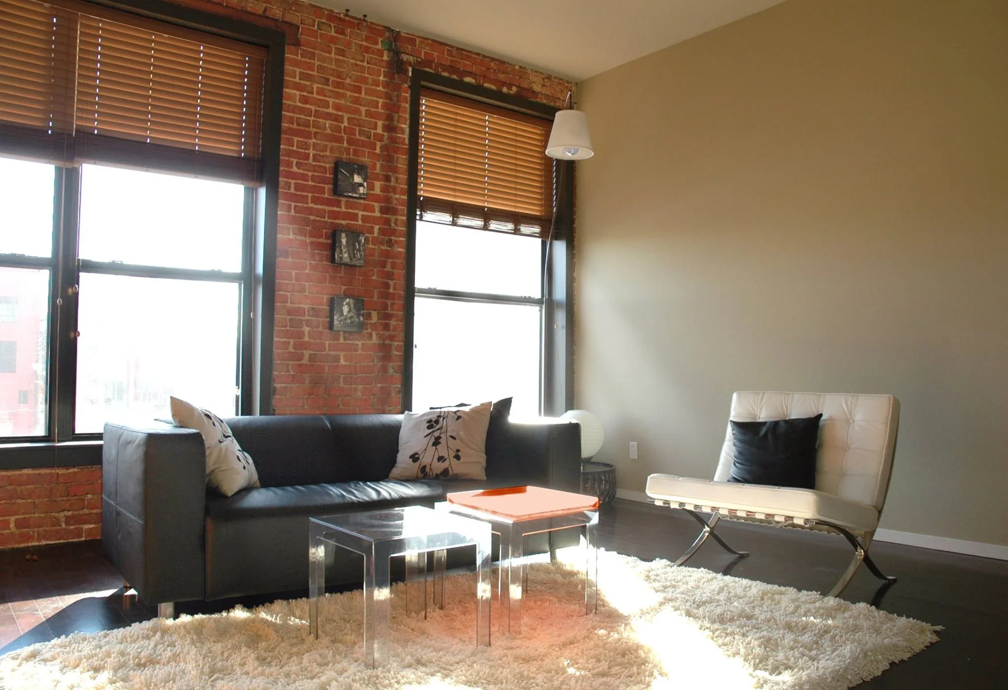 Living room with dark gray sofa, white chair with black pillow, two small clear nesting tables, a white fluffy rug, brick and beige walls, large windows with wooden blinds, and hanging white pendant light.