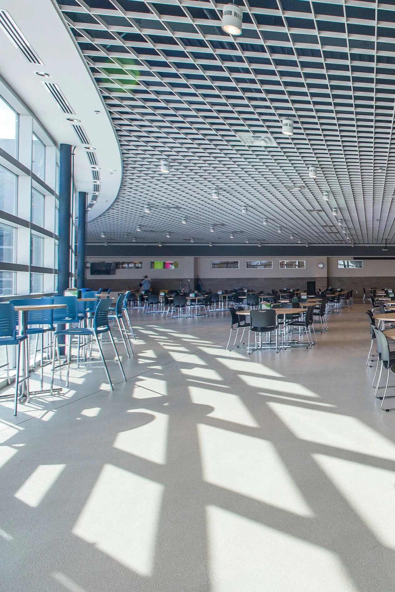 A spacious, modern interior with large windows letting in sunlight, casting geometric shadows on the floor, with empty tables and chairs arranged in an open seating area.