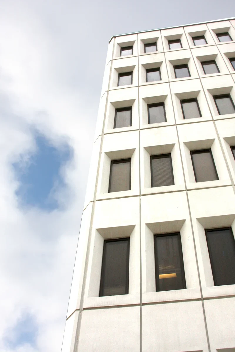 A tall white office building with multiple square windows, some with blinds, against a partly cloudy sky.