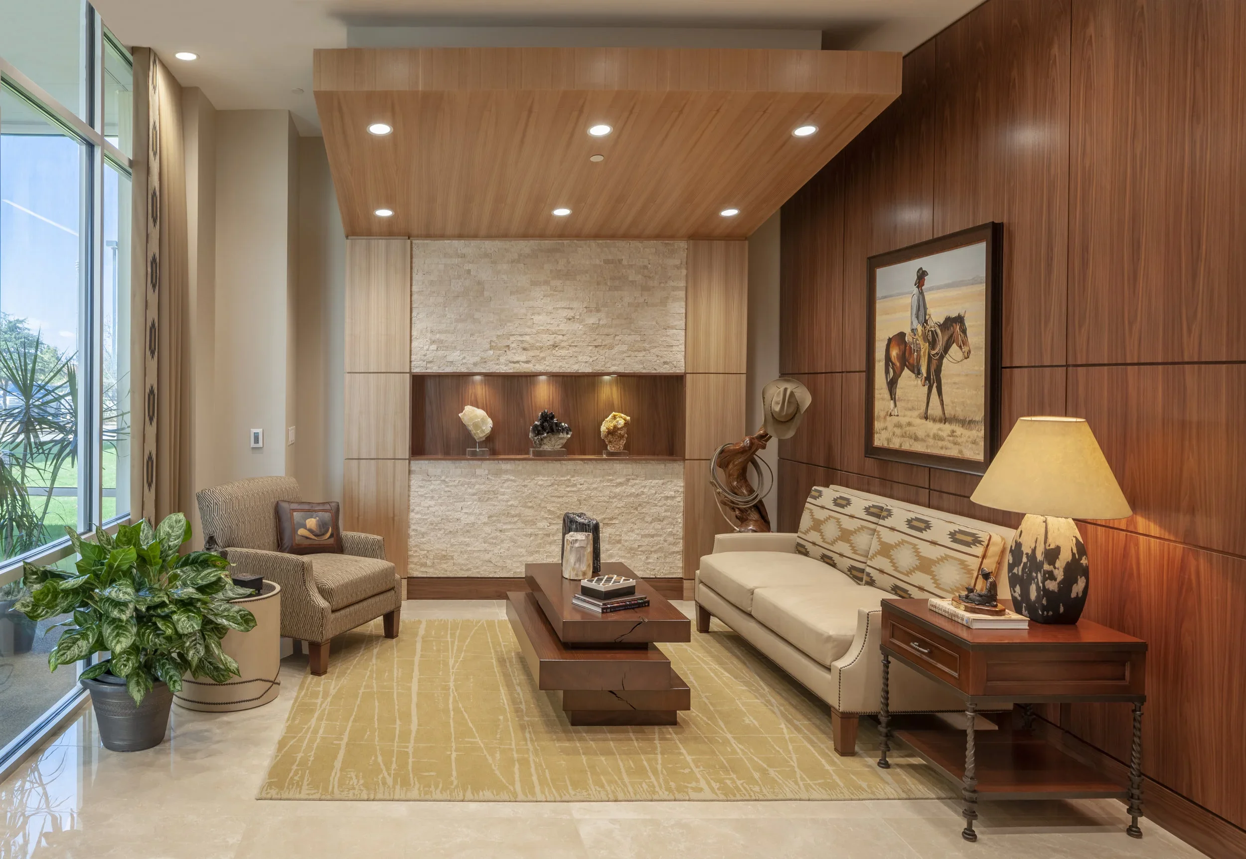 Modern lobby with a large window, beige armchair, beige sofa, wooden coffee table, side table with a lamp, artwork of a person on horseback, decorative hat on a stand, plants, display shelf with rocks, beige and cream color scheme, wood-paneled walls