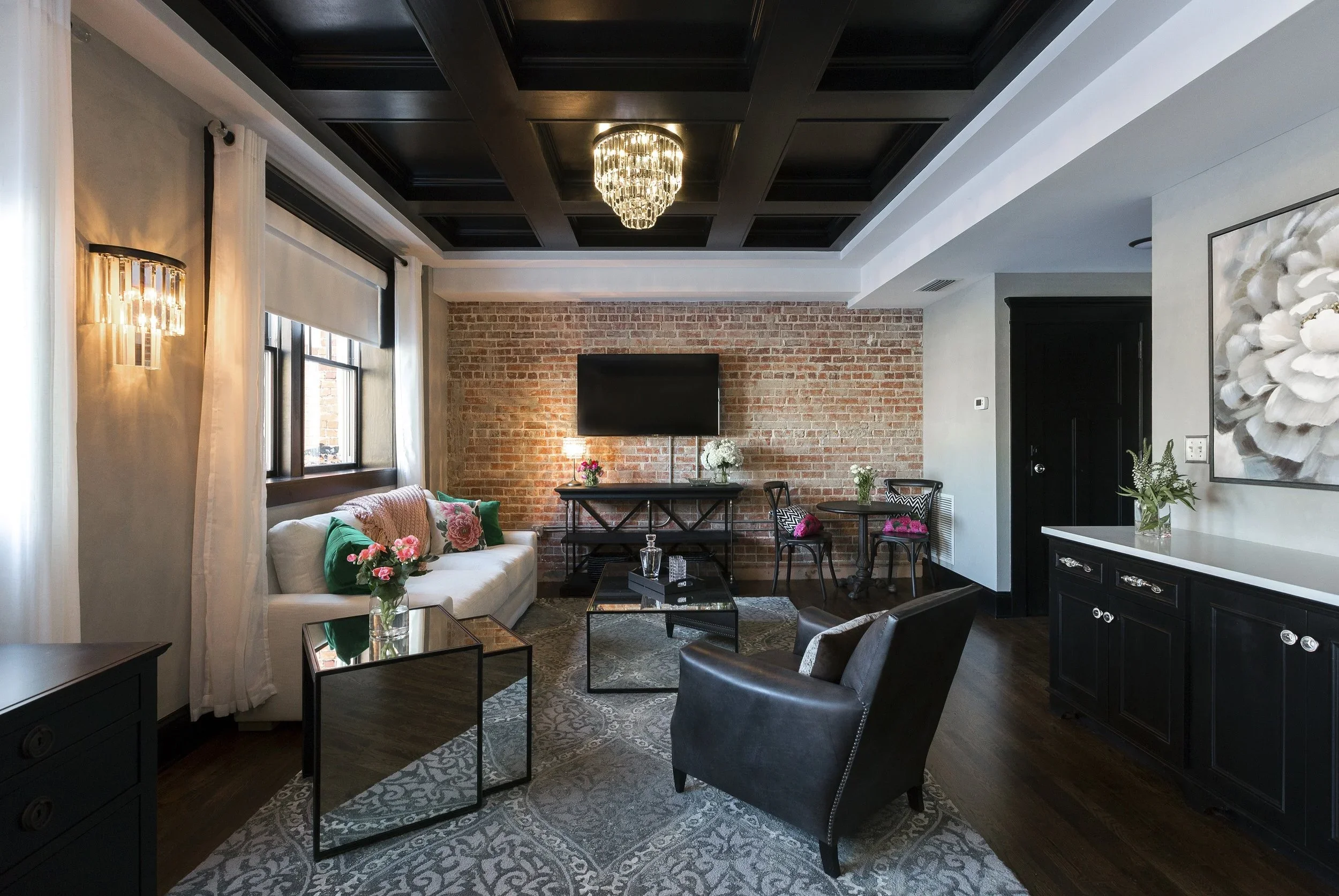 Living room with a brick accent wall, black coffered ceiling, chandelier, white sofa with floral pillows, two small glass tables, black leather armchair, dark wood flooring, wall sconces, window with white curtains, art piece, TV on brick wall, and s