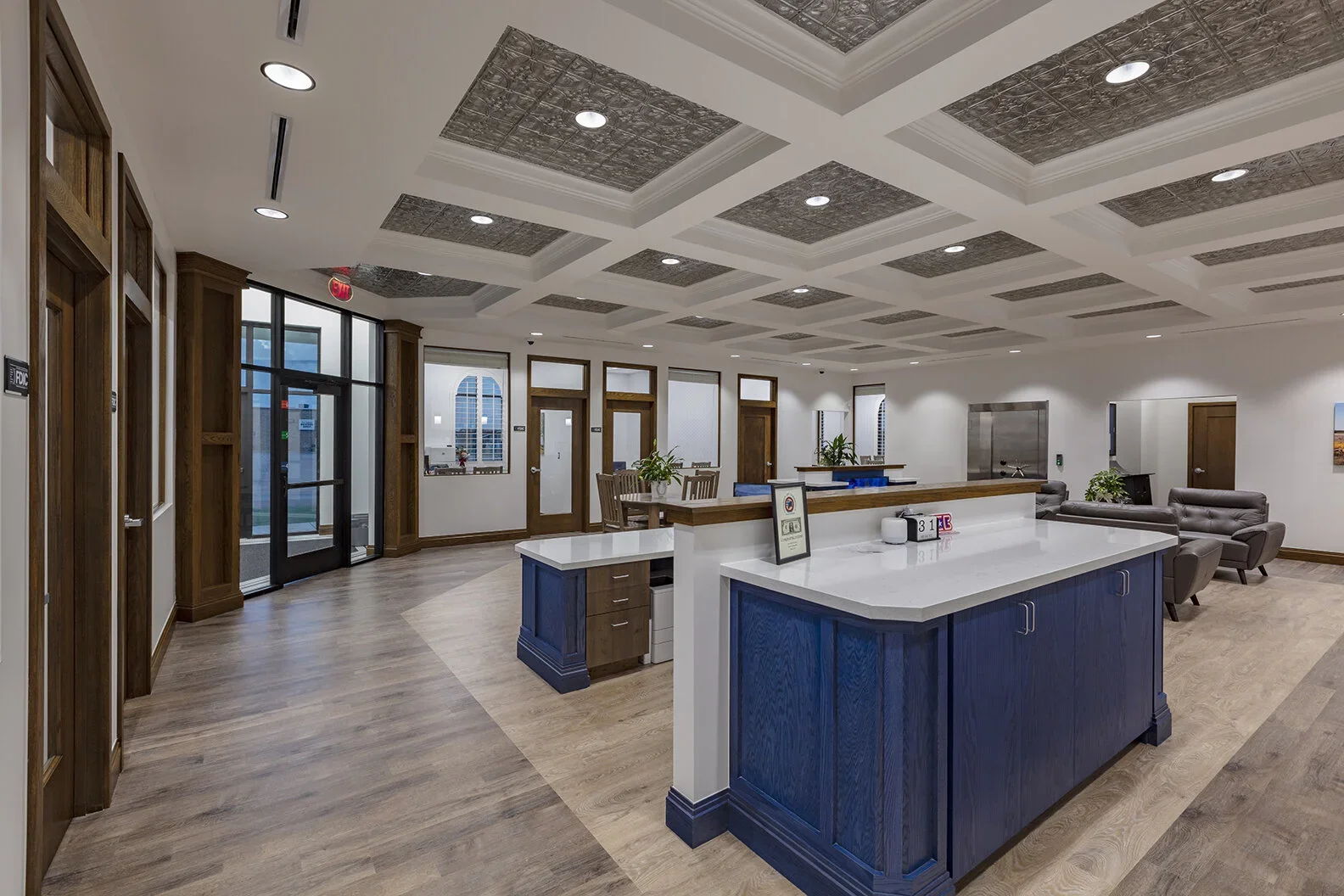 Modern waiting area with wooden flooring, blue and white reception counter, seating area with gray chairs, and decorative ceiling panels.