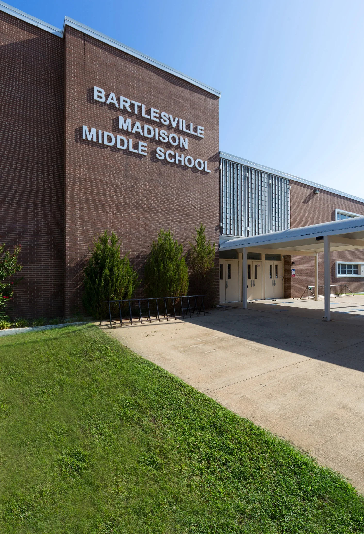 Front view of Bartlesville Madison Middle School building with brick exterior, green shrubs, sidewalk, and blue sky.