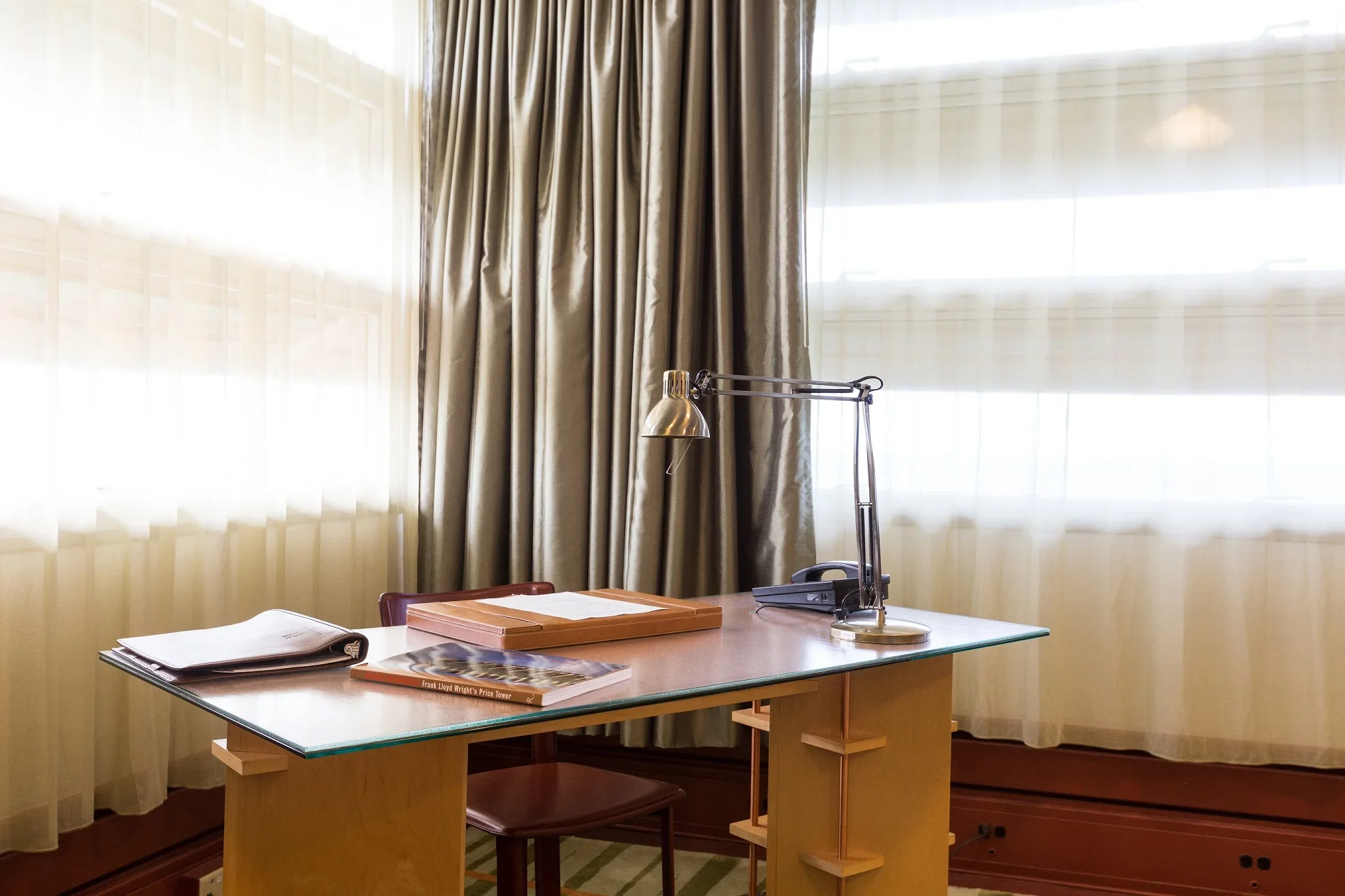 A desk with a desk lamp, a landline phone, a magazine, and documents in a room with large windows and beige curtains.