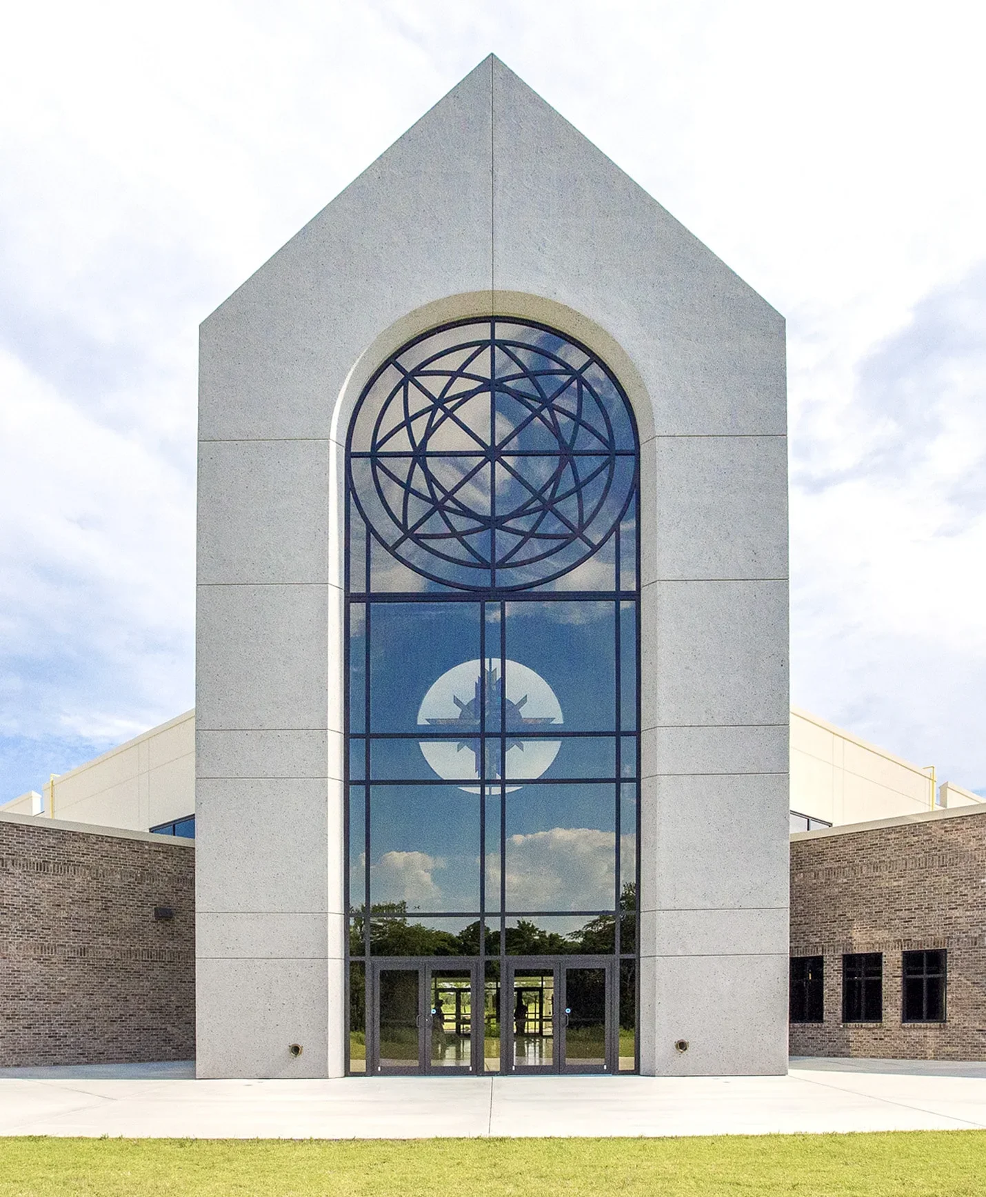 Front view of a modern church building with a large arched stained glass window featuring geometric patterns, surrounded by brick and concrete walls, and a grassy area in front.