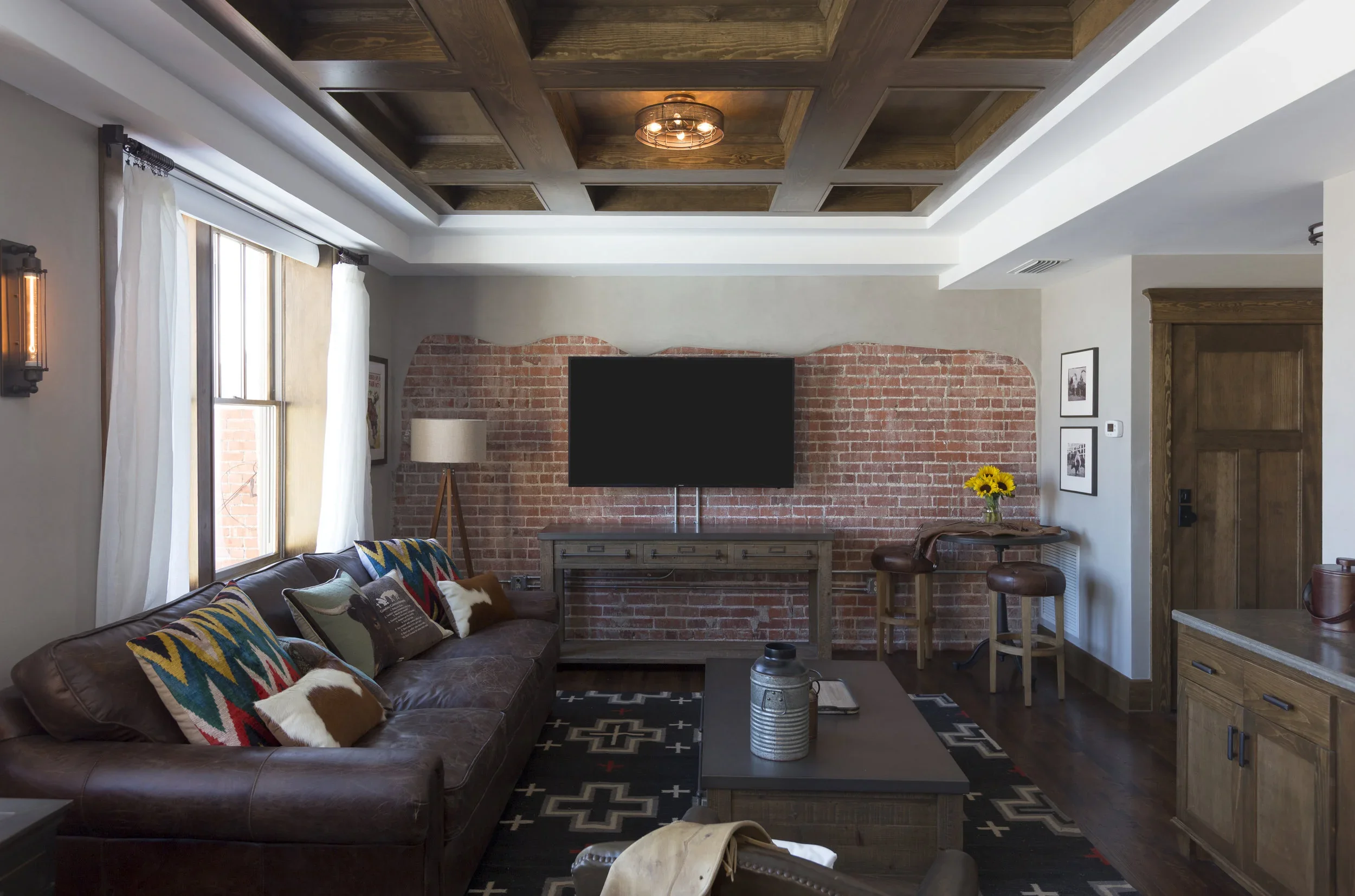 A cozy living room with a leather sofa, colorful pillows, a wooden coffee table, a large flat-screen TV mounted on an exposed brick wall, a floor lamp, a window with white curtains, a small table with sunflowers, and a wooden cabinet.