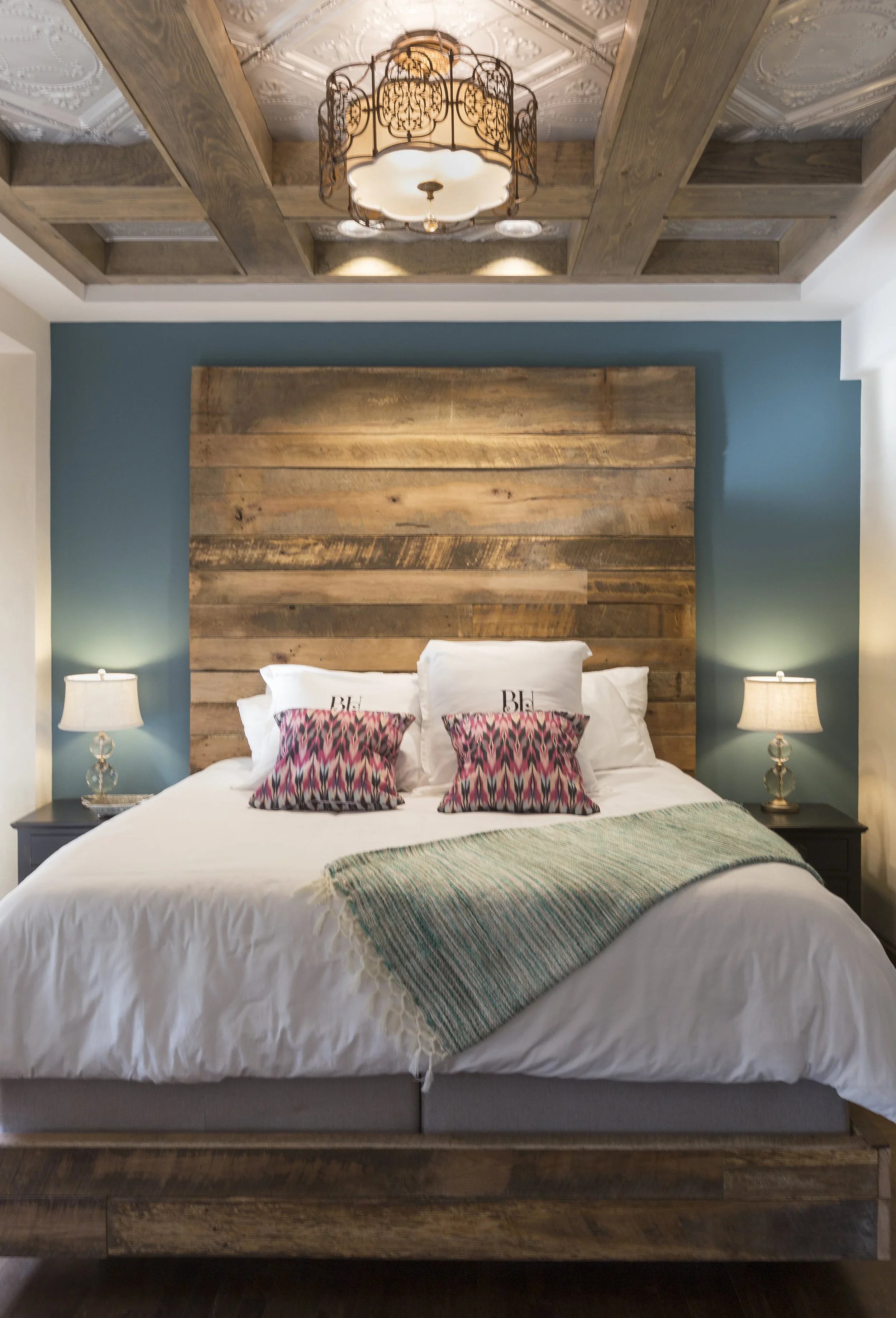 A bedroom with a large bed featuring white bedding, decorative pillows, and a wooden headboard. The room has teal walls, two bedside tables with lamps, and a wooden and glass ceiling fixture.